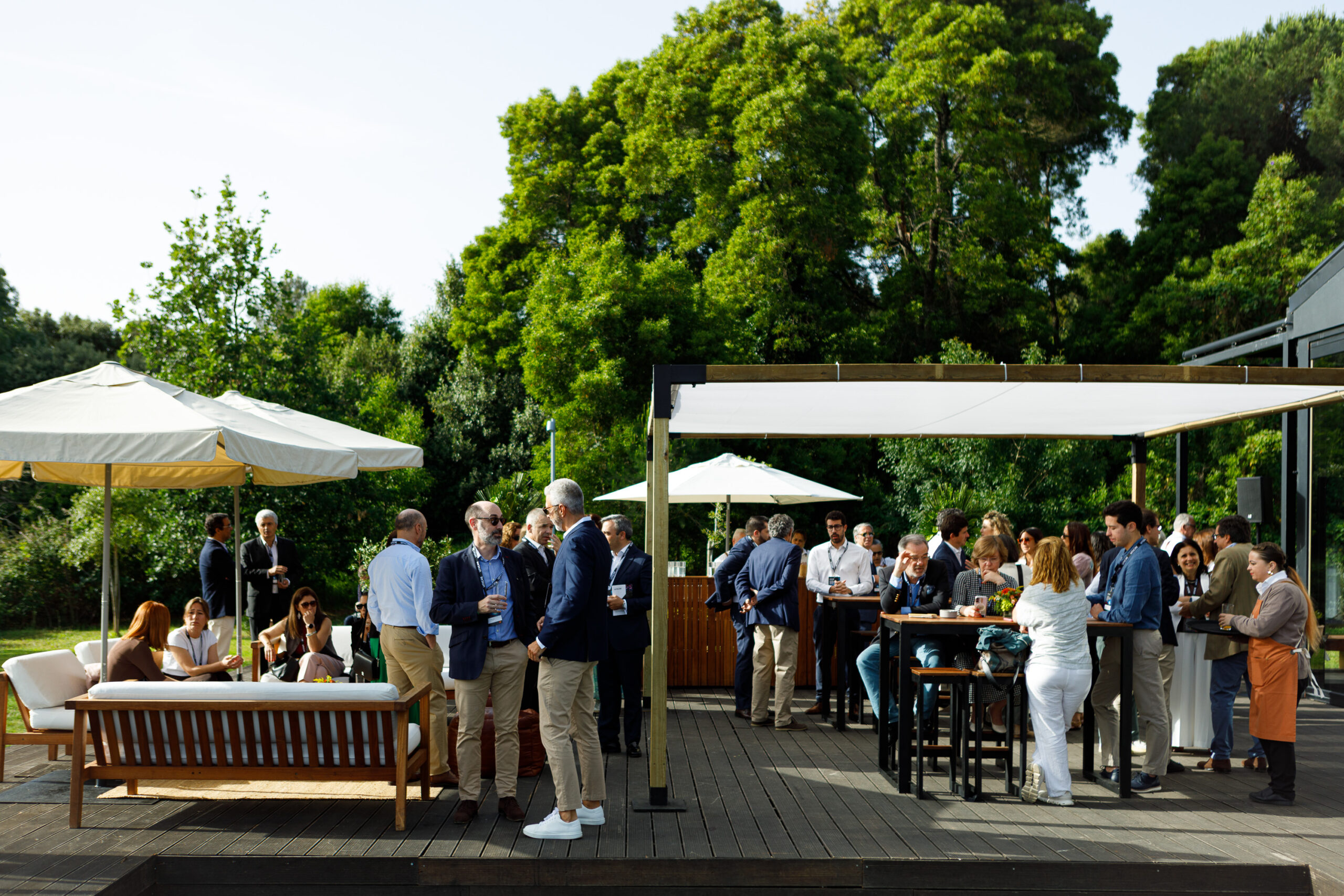 Outdoor corporate networking event with groups of professionals chatting under white tents and umbrellas on a wooden deck surrounded by trees.