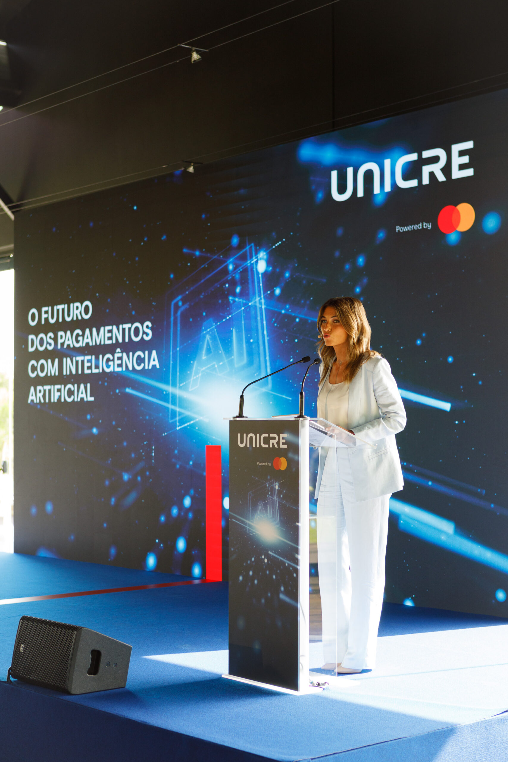 Woman in a light suit giving a speech at a UNICRE stage podium, with a blue digital backdrop and Mastercard logo visible