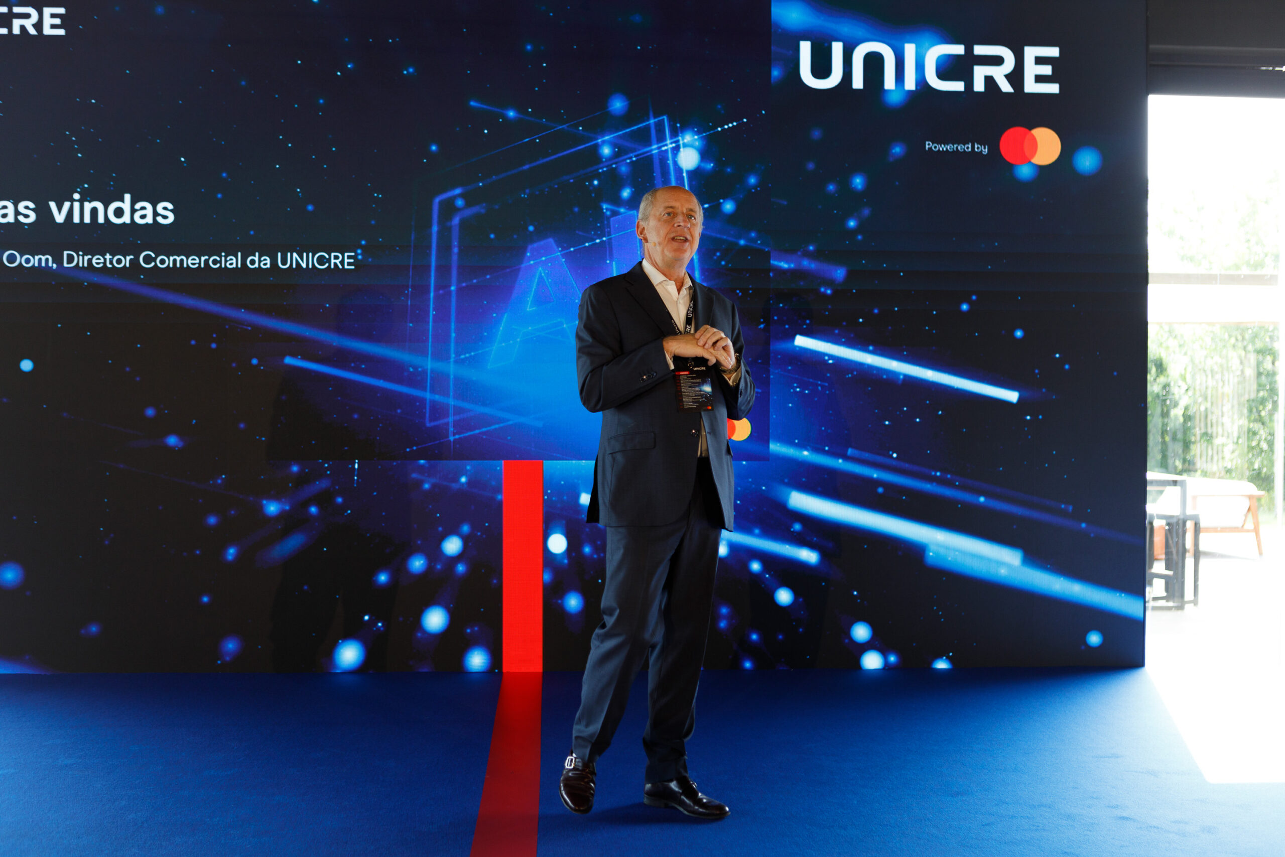 Male presenter in a navy suit speaking on a blue tech-themed stage with UNICRE branding behind him.