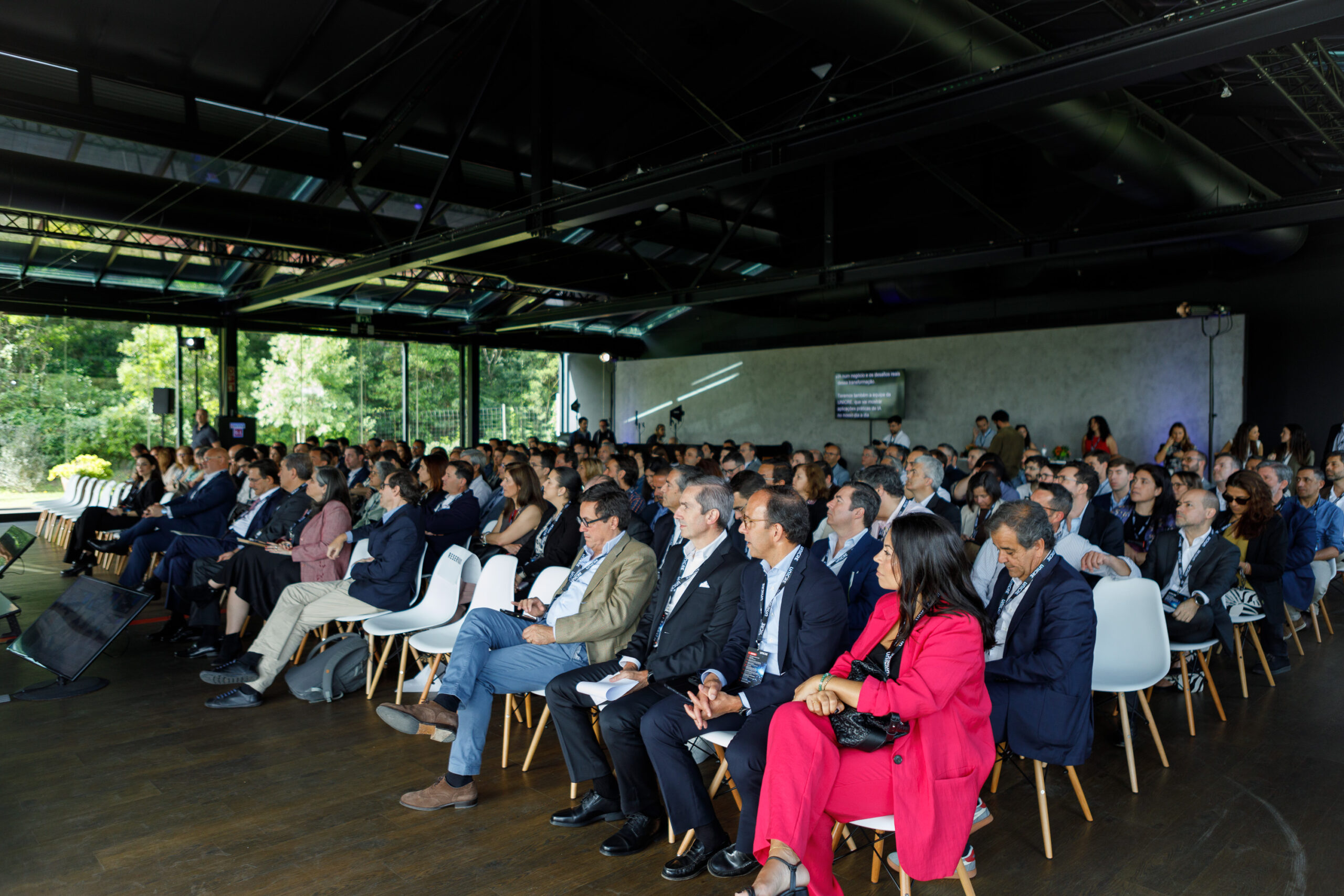 Professional audience seated in rows at a daytime conference in a modern venue, with a stage and screens in the background