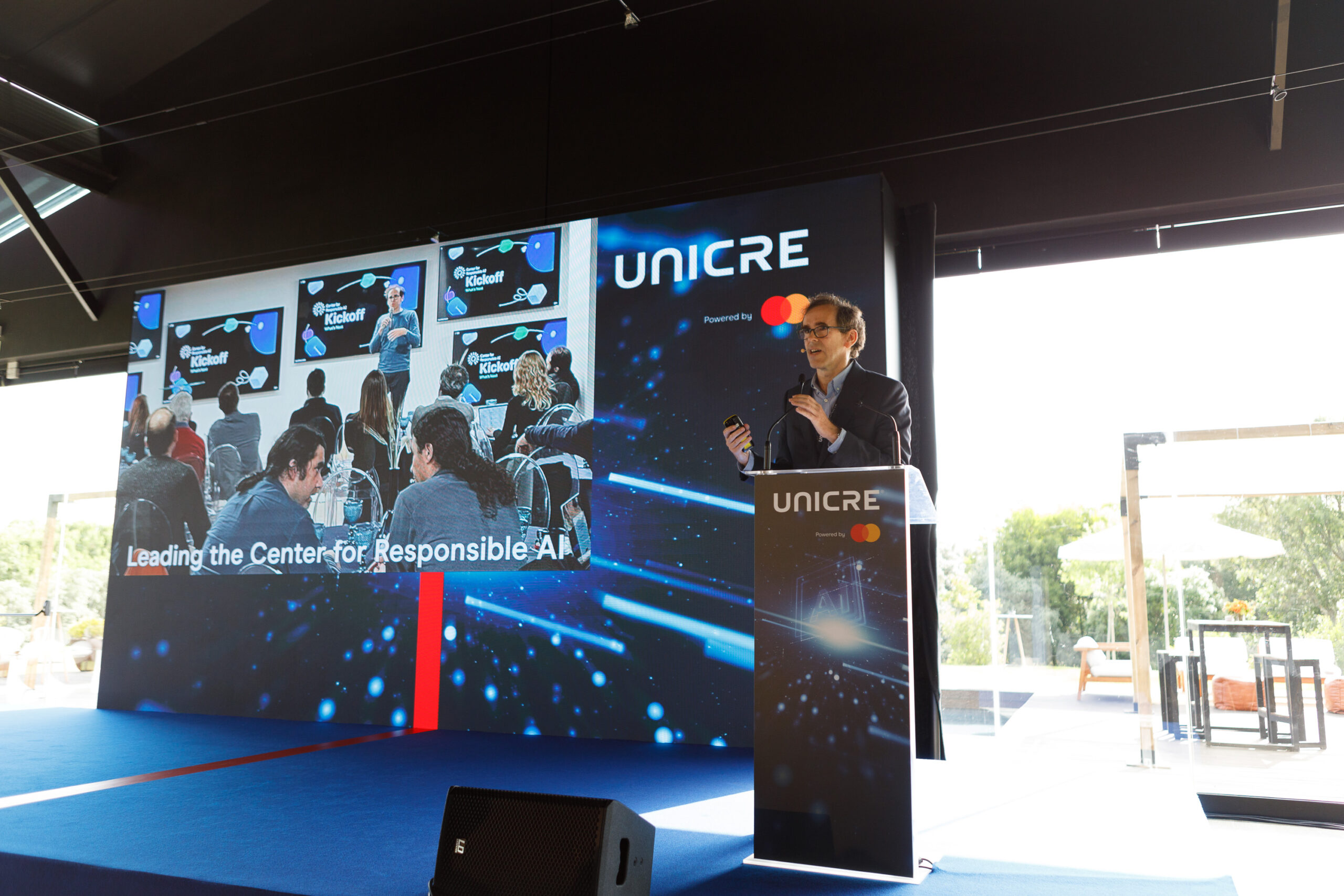 Speaker at a podium giving a presentation on a stage with a large screen showing an audience and the UNICRE branding above and to the side.