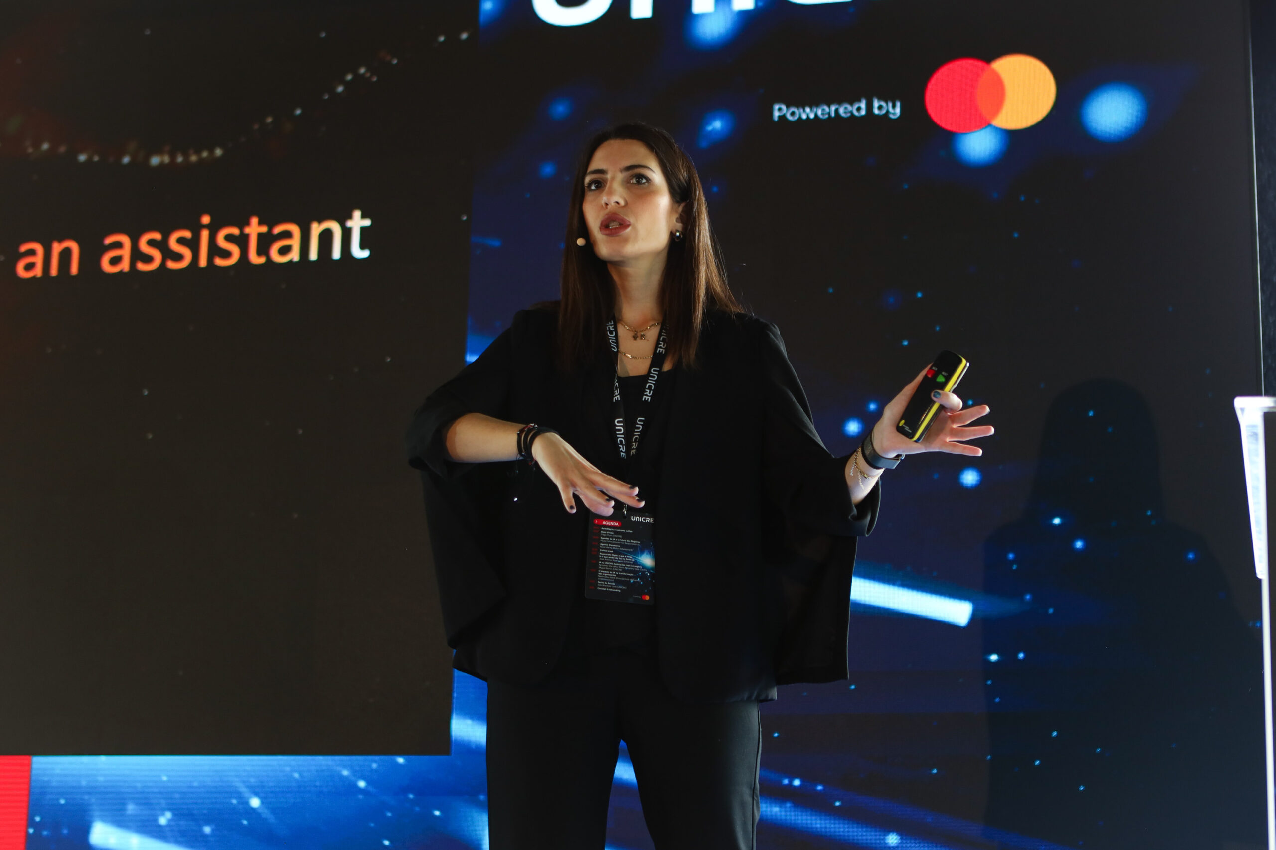 Female presenter on a dark stage, gesturing with hands, wearing a black outfit and lanyard, holding a yellow-and-black device against a tech-themed backdrop.