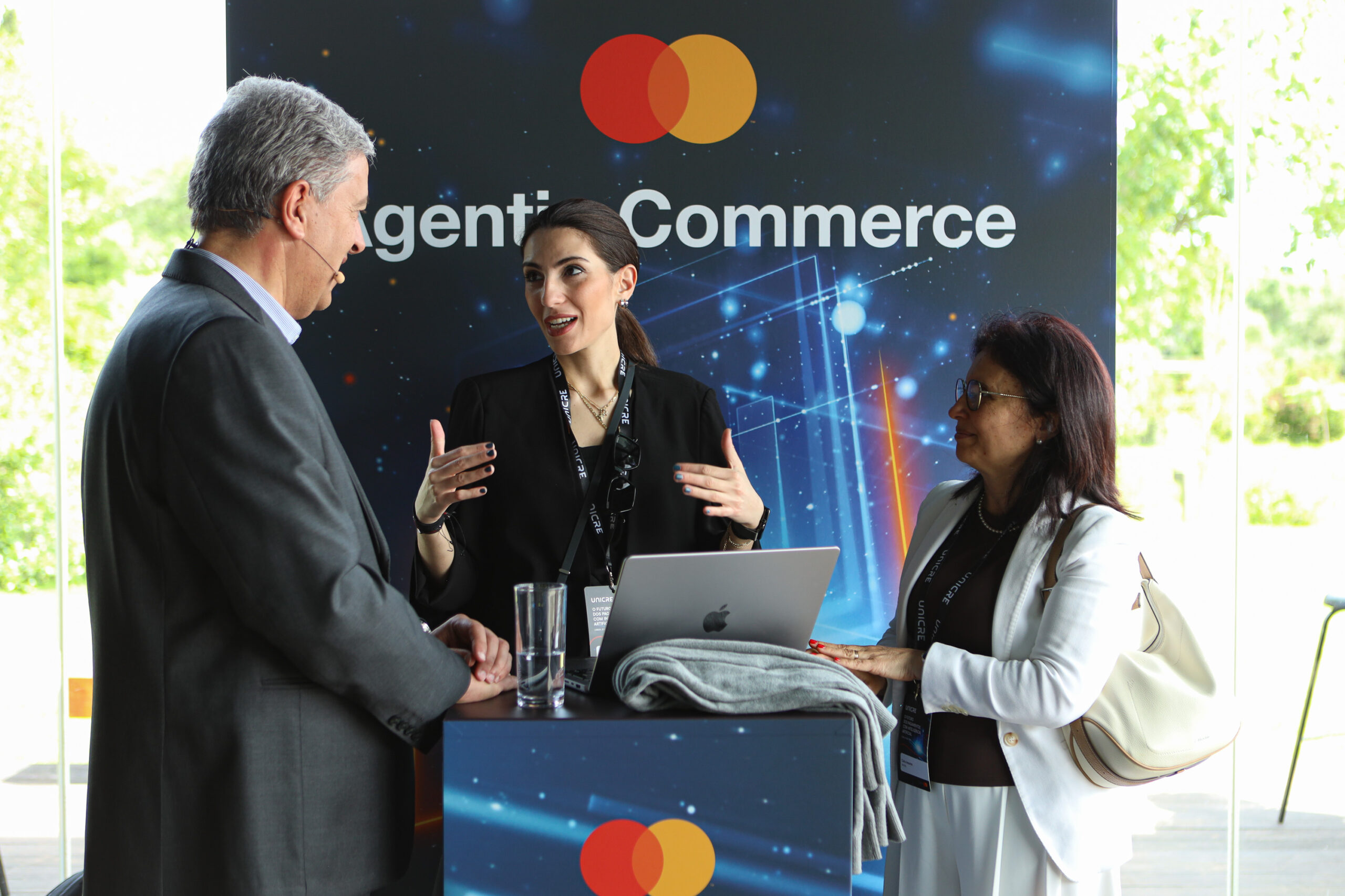 Three professionals discuss at a Mastercard-branded event booth with a laptop on the counter.