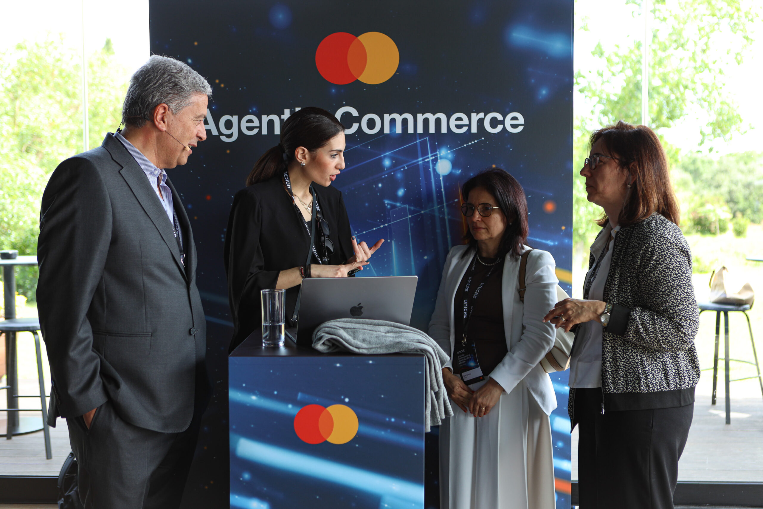 Four professionals in business attire stand around a counter at an event booth with a Mastercard branded backdrop, engaging in discussion.