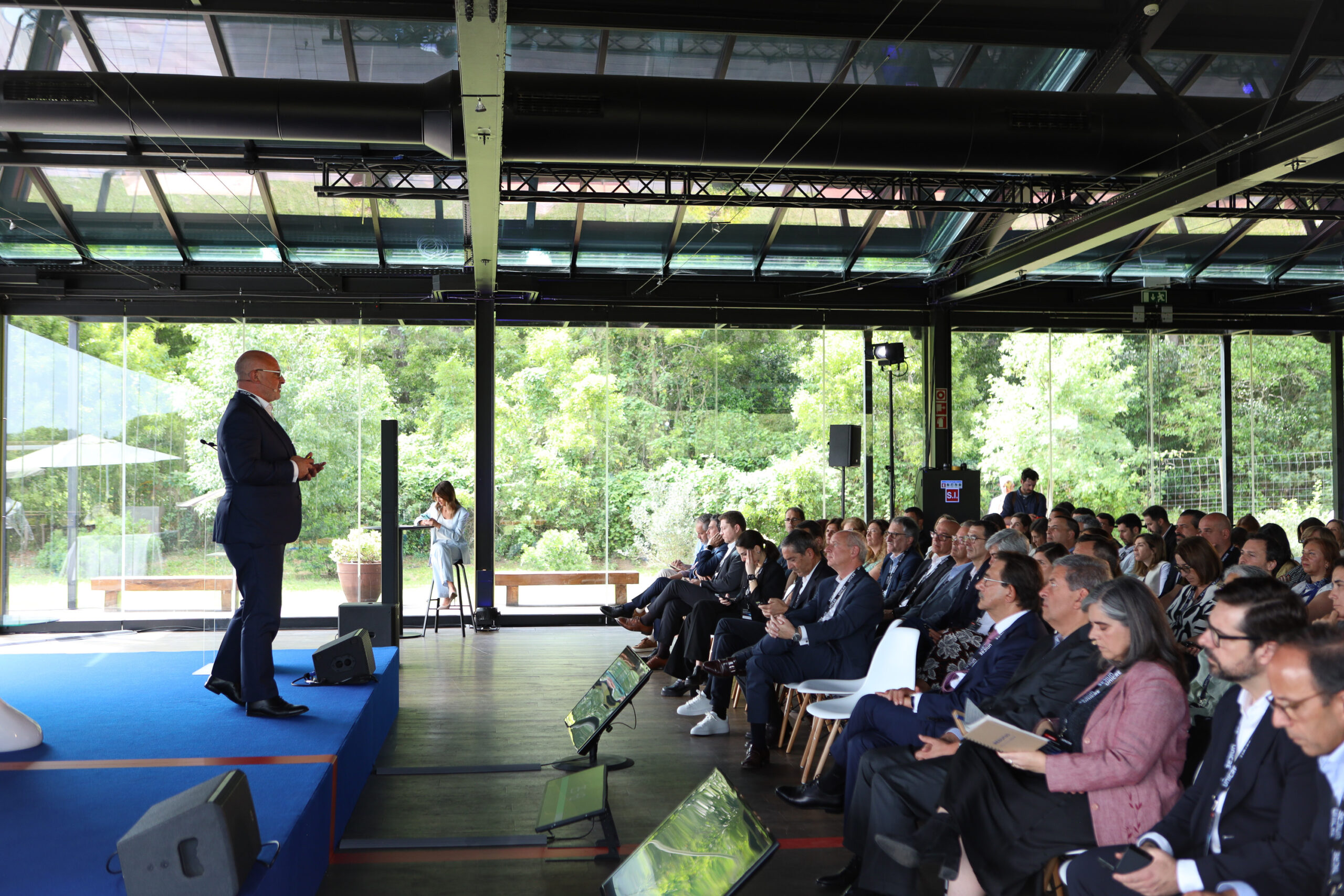 Man in a navy suit giving a talk on a blue stage to an attentive audience in a glass-walled conference hall with greenery outside.