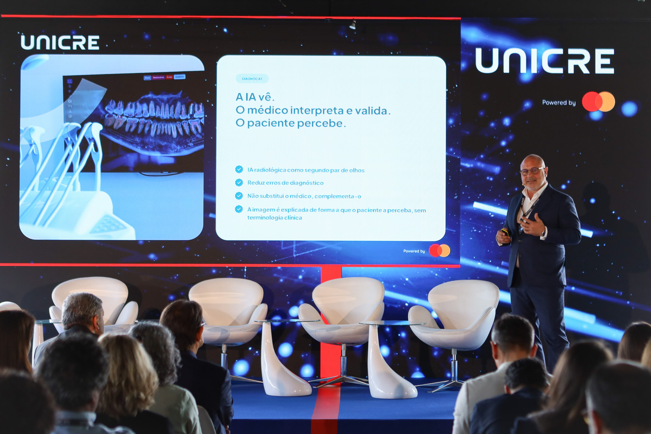 Presenter in a suit on a stage beside a large screen showing UNICRE branding and a dental image; audience seated below.