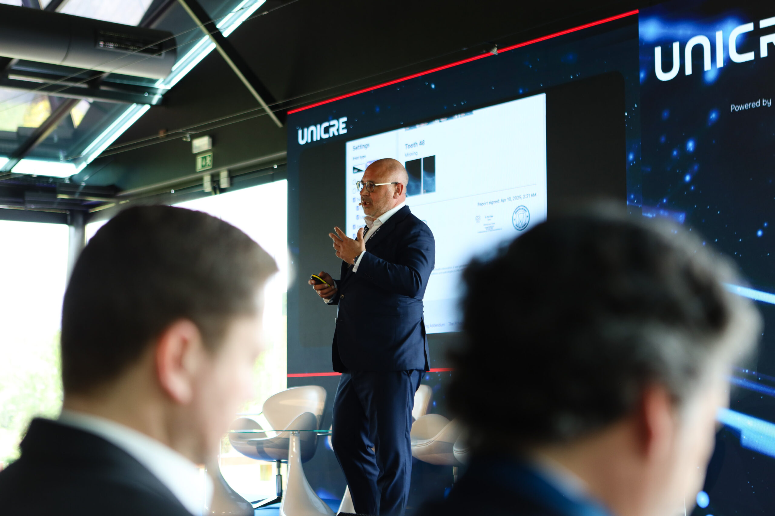 Male presenter in a suit gesturing while speaking to an audience in a modern conference room; large screen shows slides with UNICRE branding behind him.