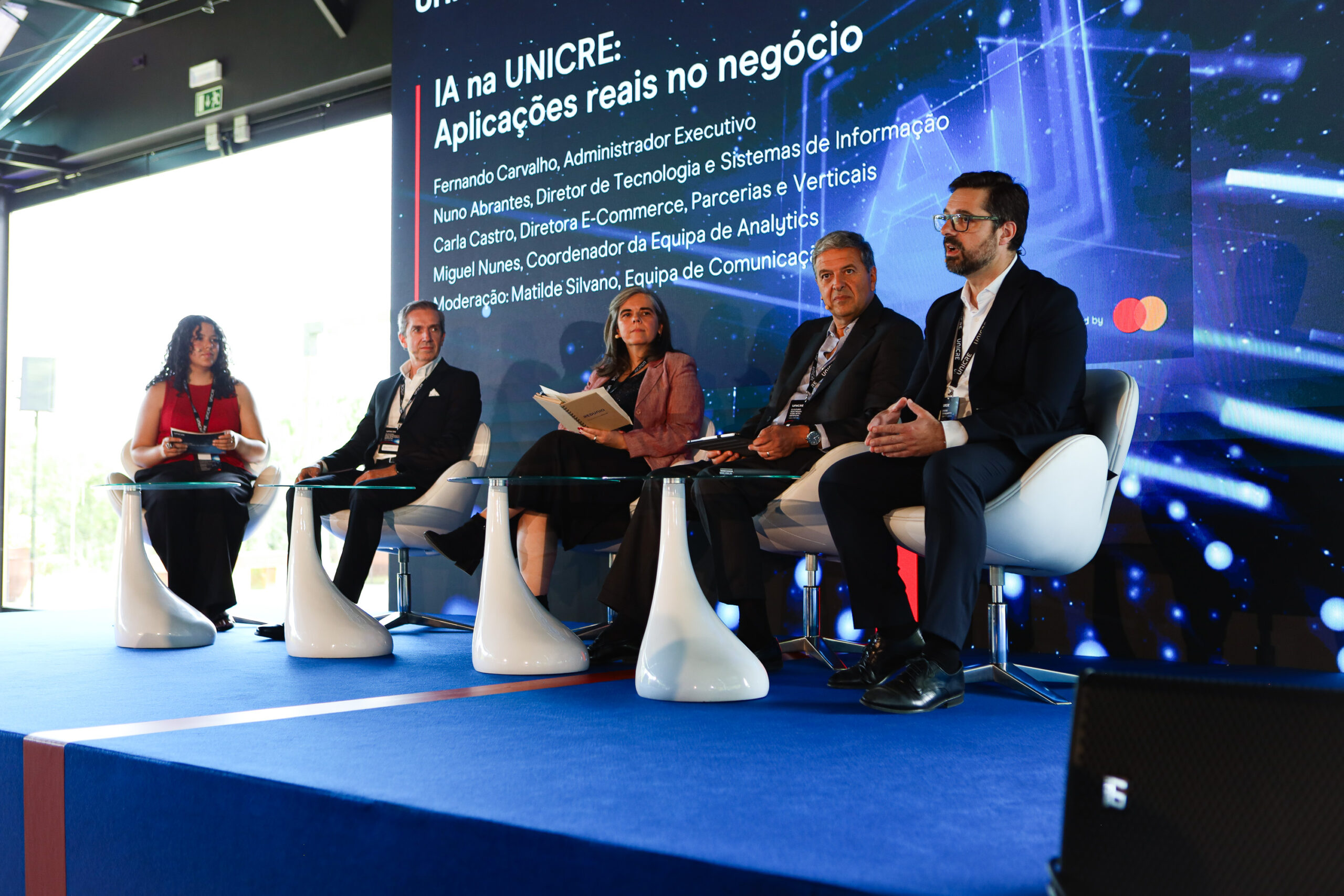 Panel of five speakers seated on a stage during a conference, with a large blue screen showing text behind them.