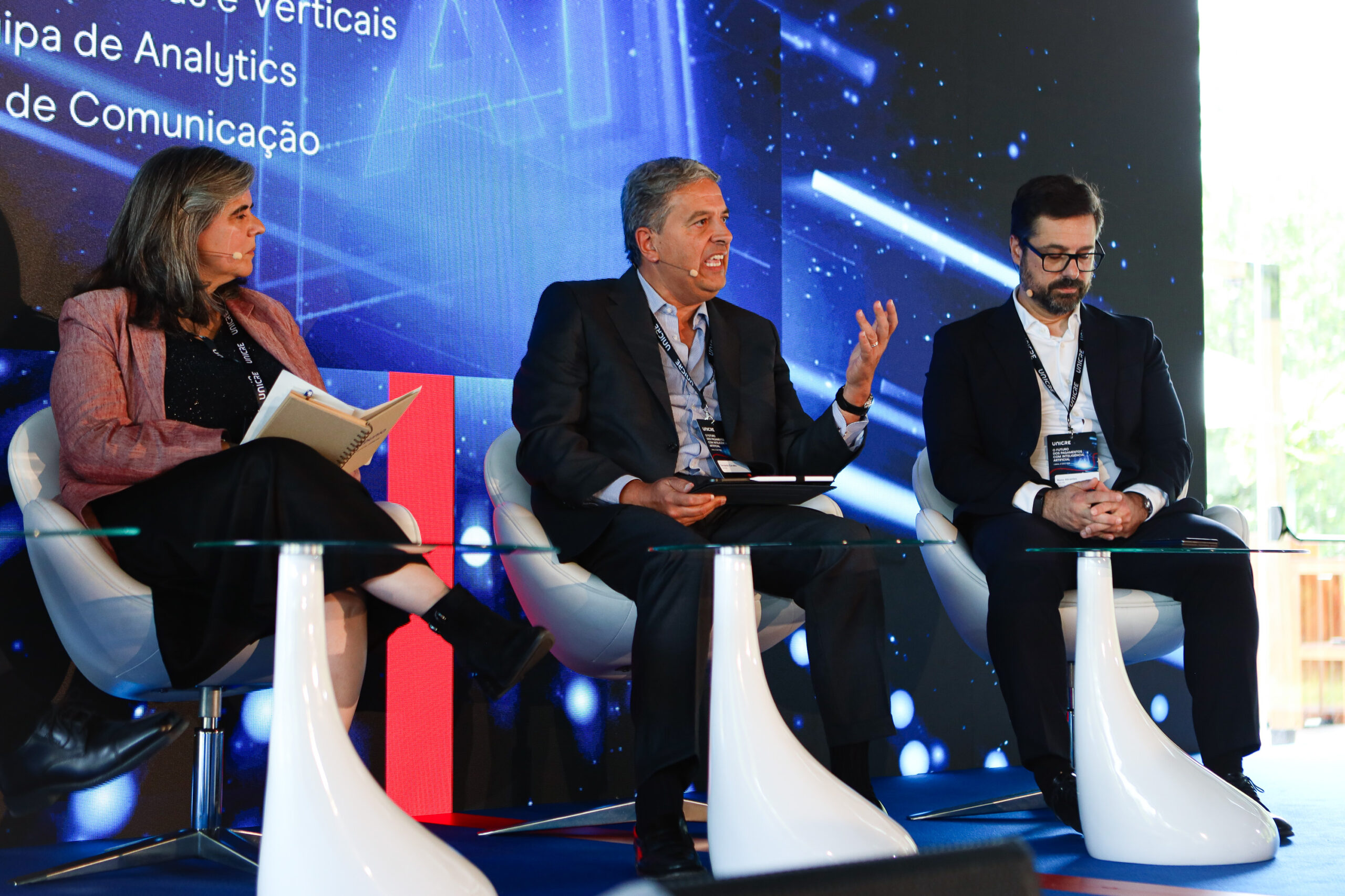 Three panelists seated on a stage in front of a blue, high-tech backdrop; the middle man speaks into a headset mic while others listen, one holds a notebook.