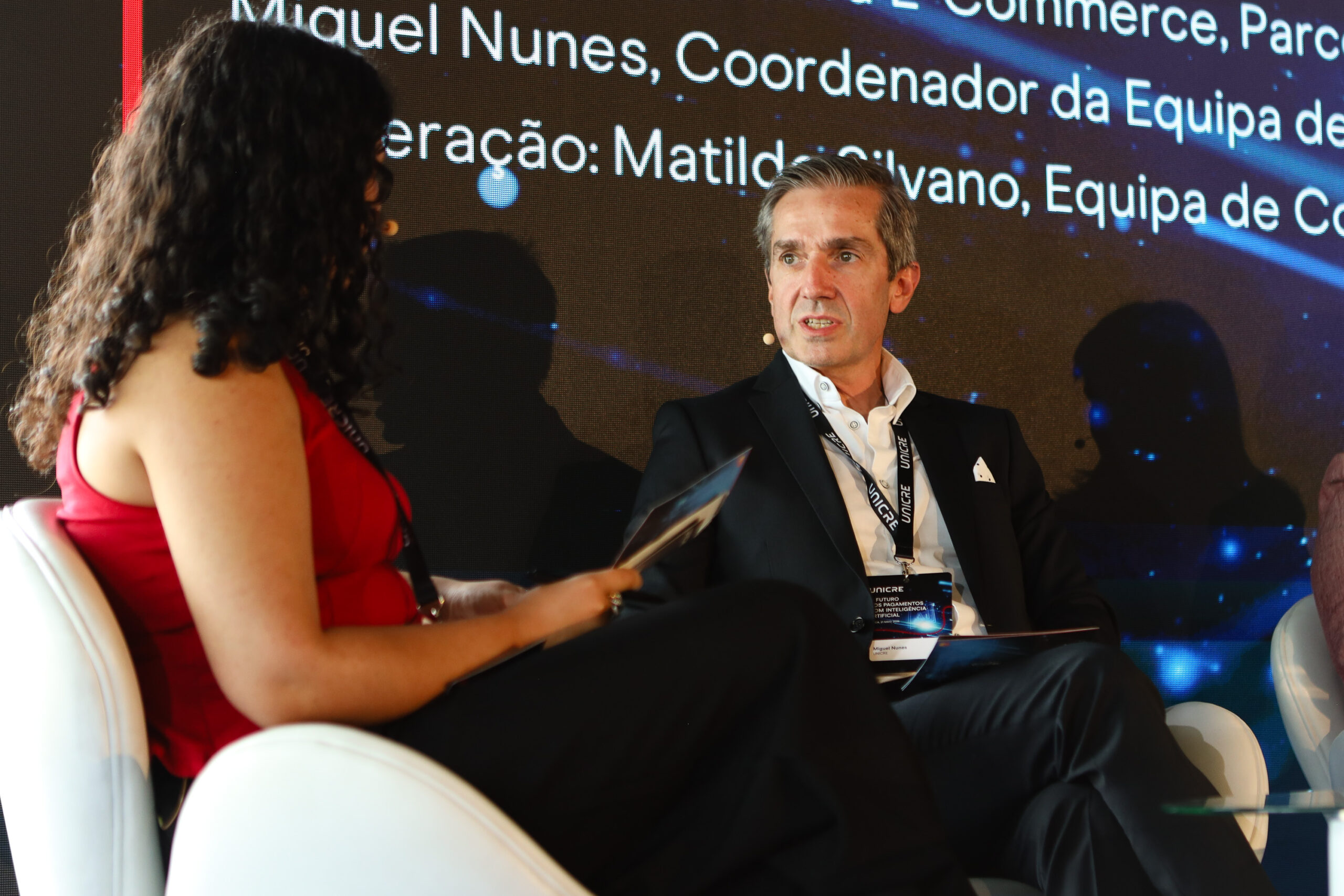 Two people seated facing each other on a stage; a woman in a red dress speaks while a man in a dark suit listens, with event badges and a large screen backdrop behind them.