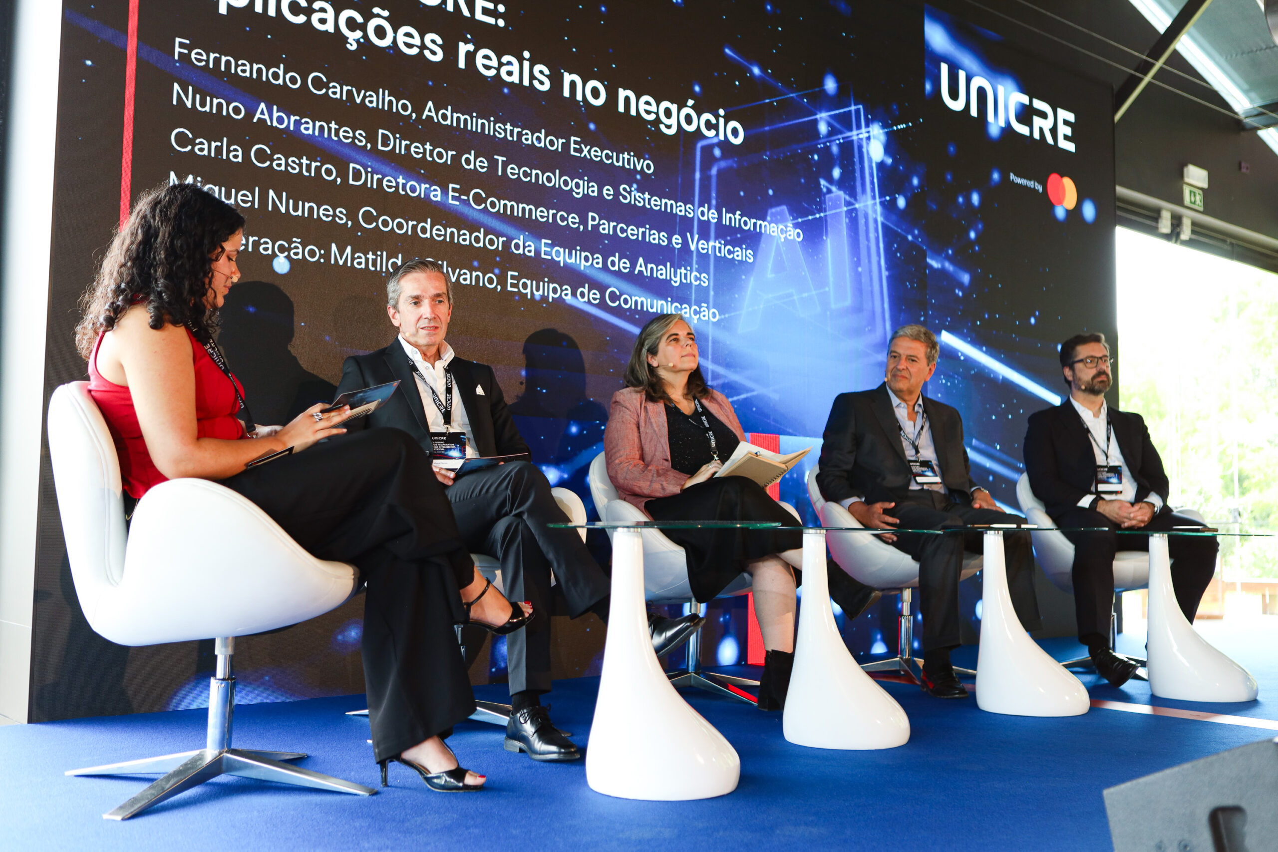 Panel of five professionals seated on a stage with a blue carpet, a large digital screen behind them displaying Portuguese text and names (conference panel).