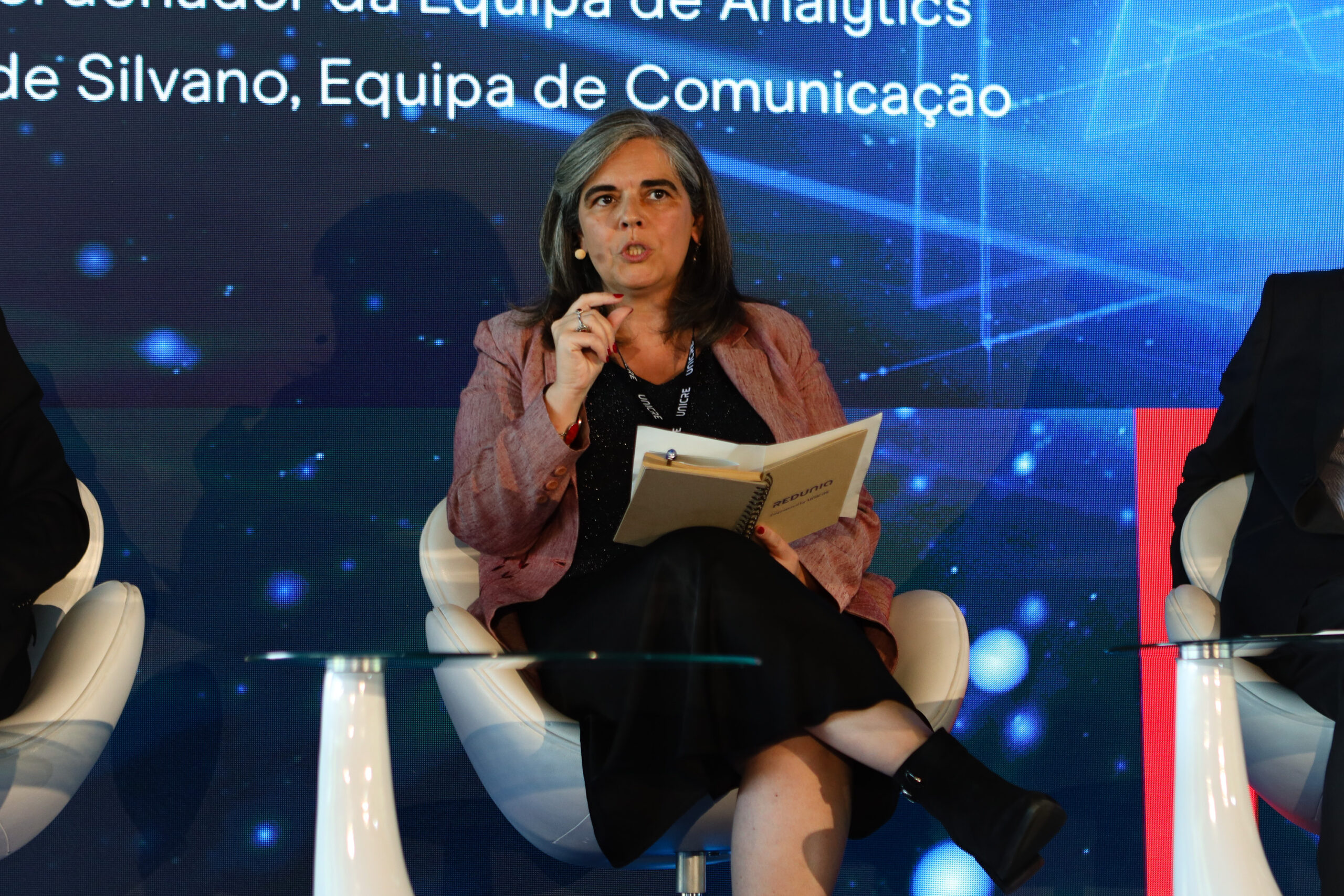 Woman panelist speaking into a headset mic, holding a notebook, seated on a white chair with a blue tech backdrop behind her.