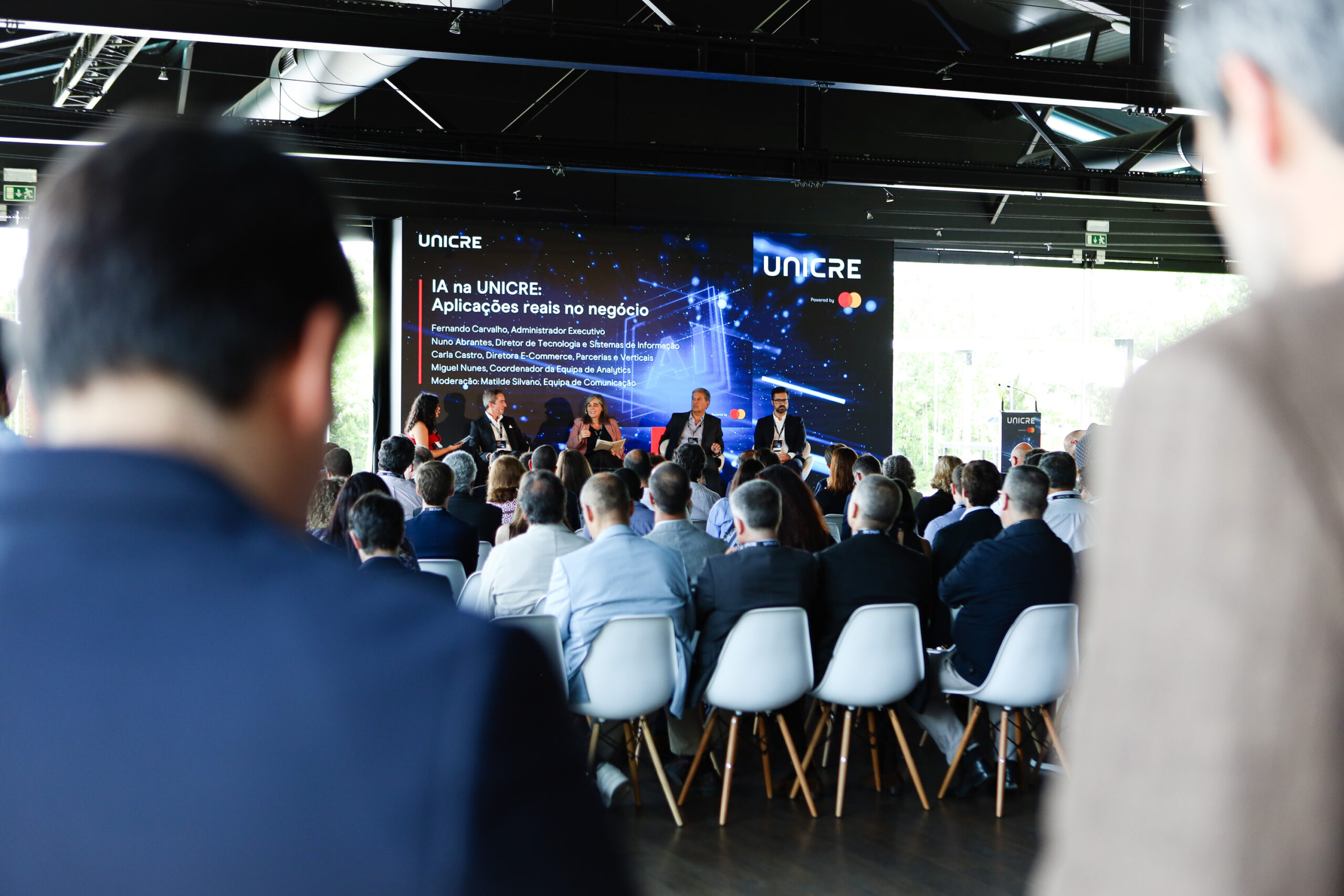 Panel discussion on a stage with a blue UNICRE banner and audience in a conference hall behind the foreground blur.
