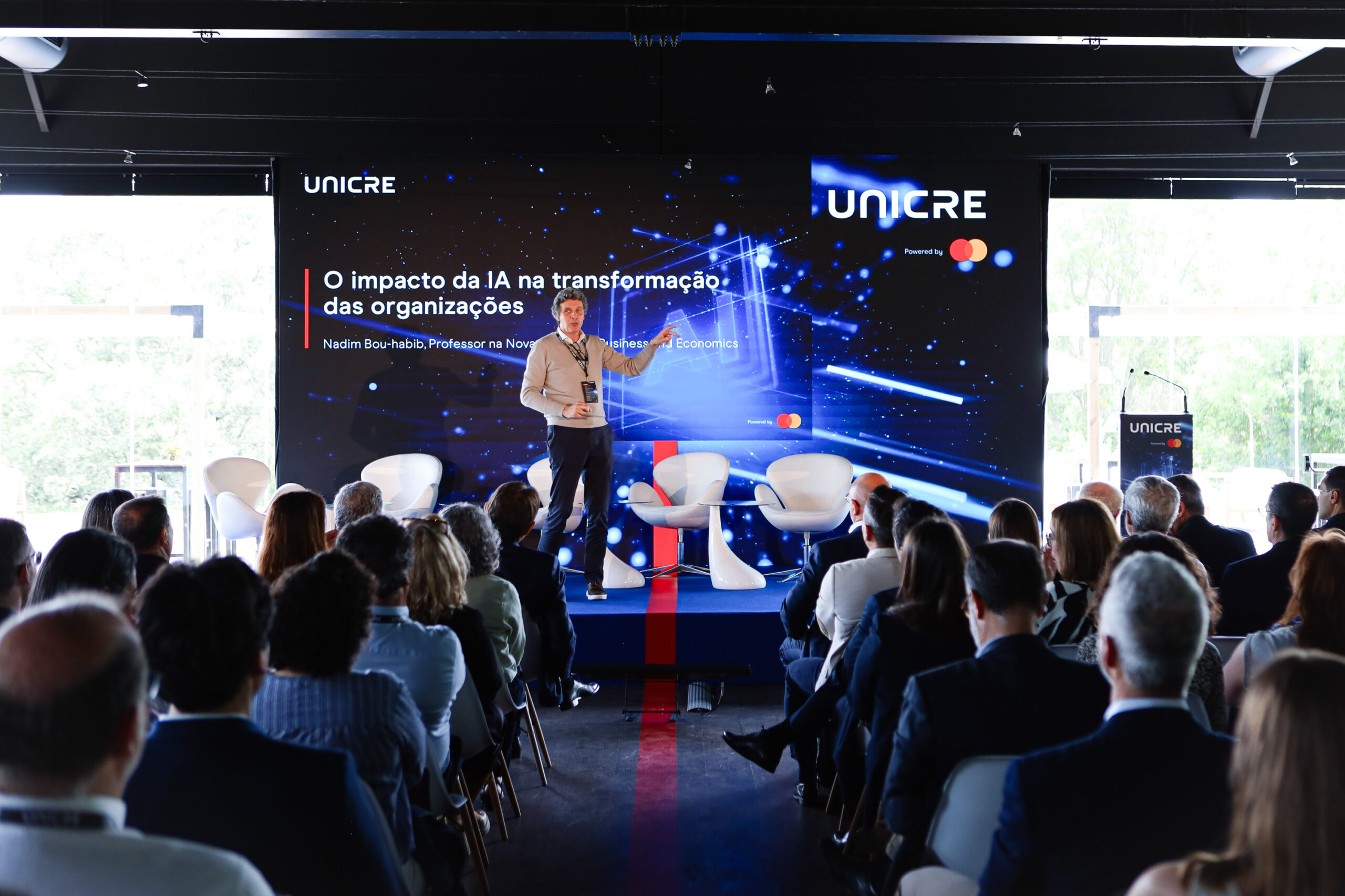 Presenter on a stage giving a talk in a conference hall with UNICRE branding and an audience seated before a large blue, tech-themed backdrop.