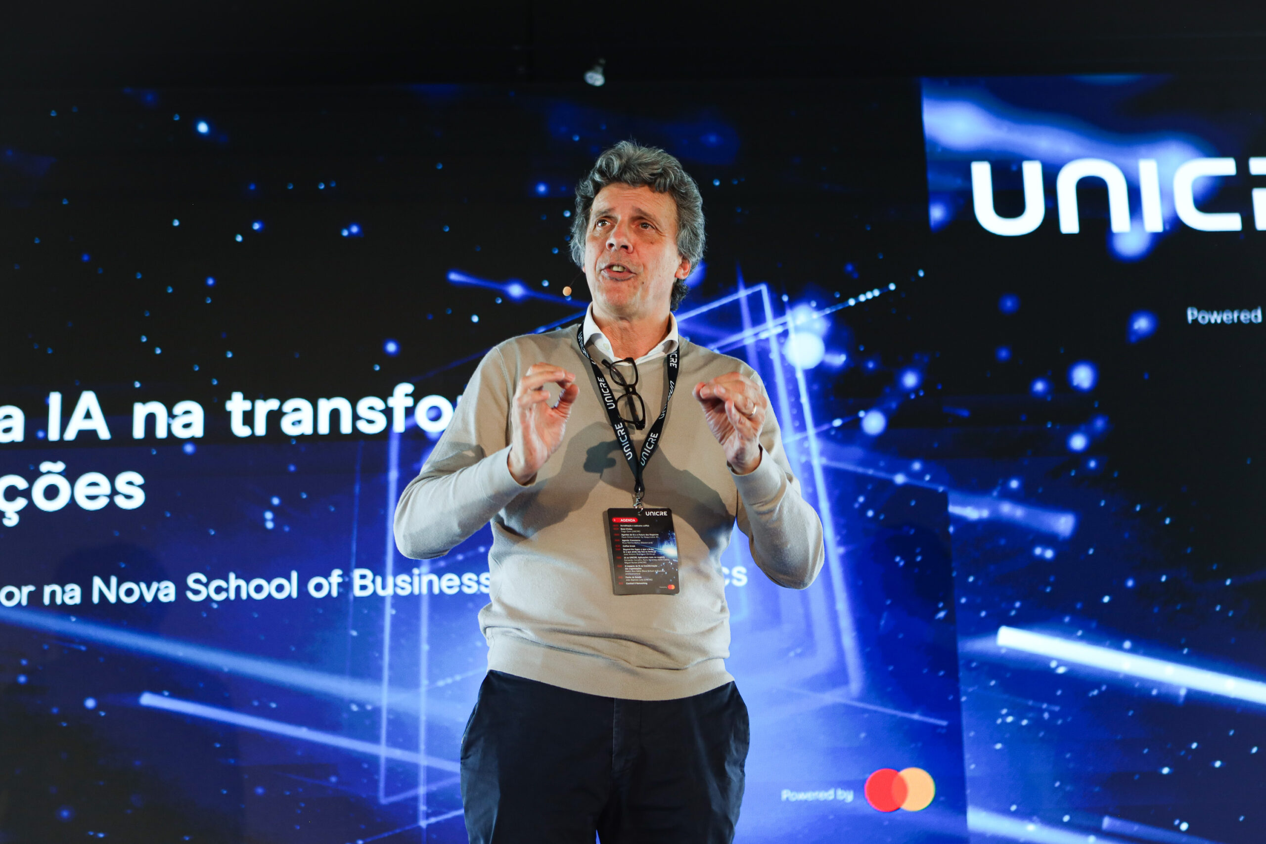 Male speaker on stage delivering a talk at a tech conference, wearing a lanyard and badge with a blue digital backdrop behind him.