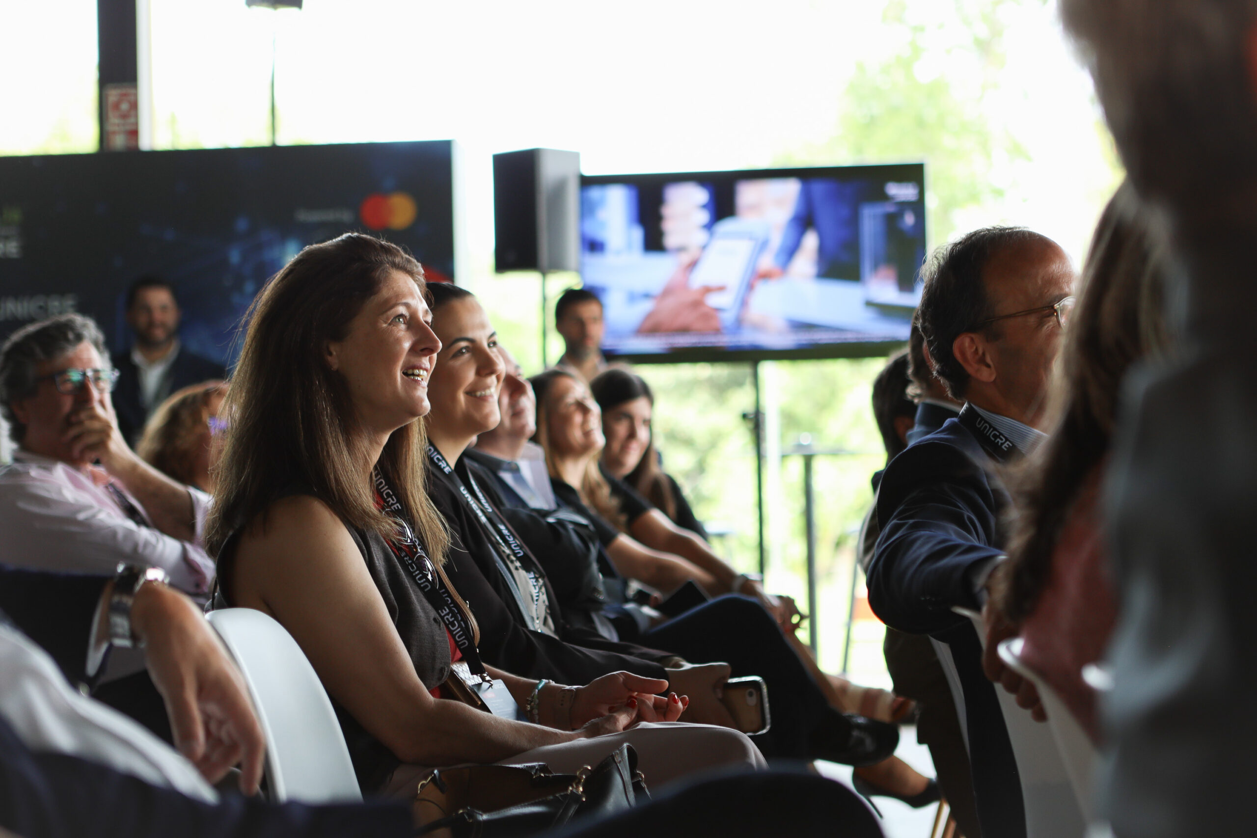 Smiling conference attendees seated in rows, wearing lanyards, watching a presentation on large screens in the background.
