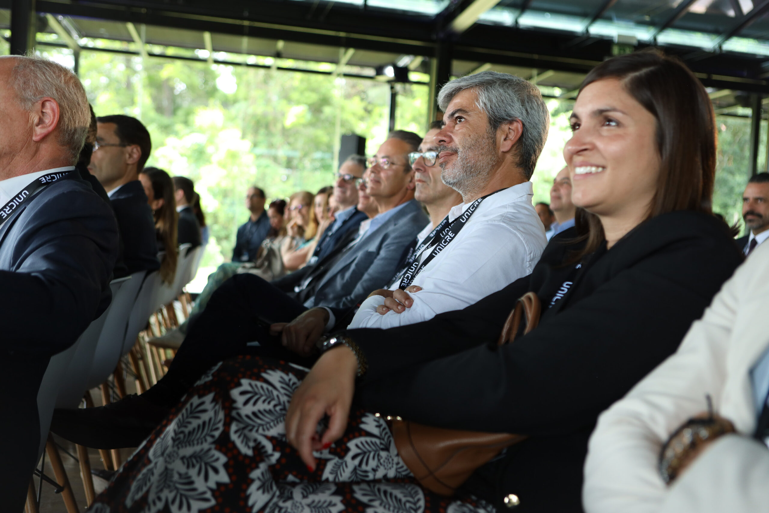 Group of professionally dressed attendees seated in a bright indoor venue, listening to a presentation or speaker.