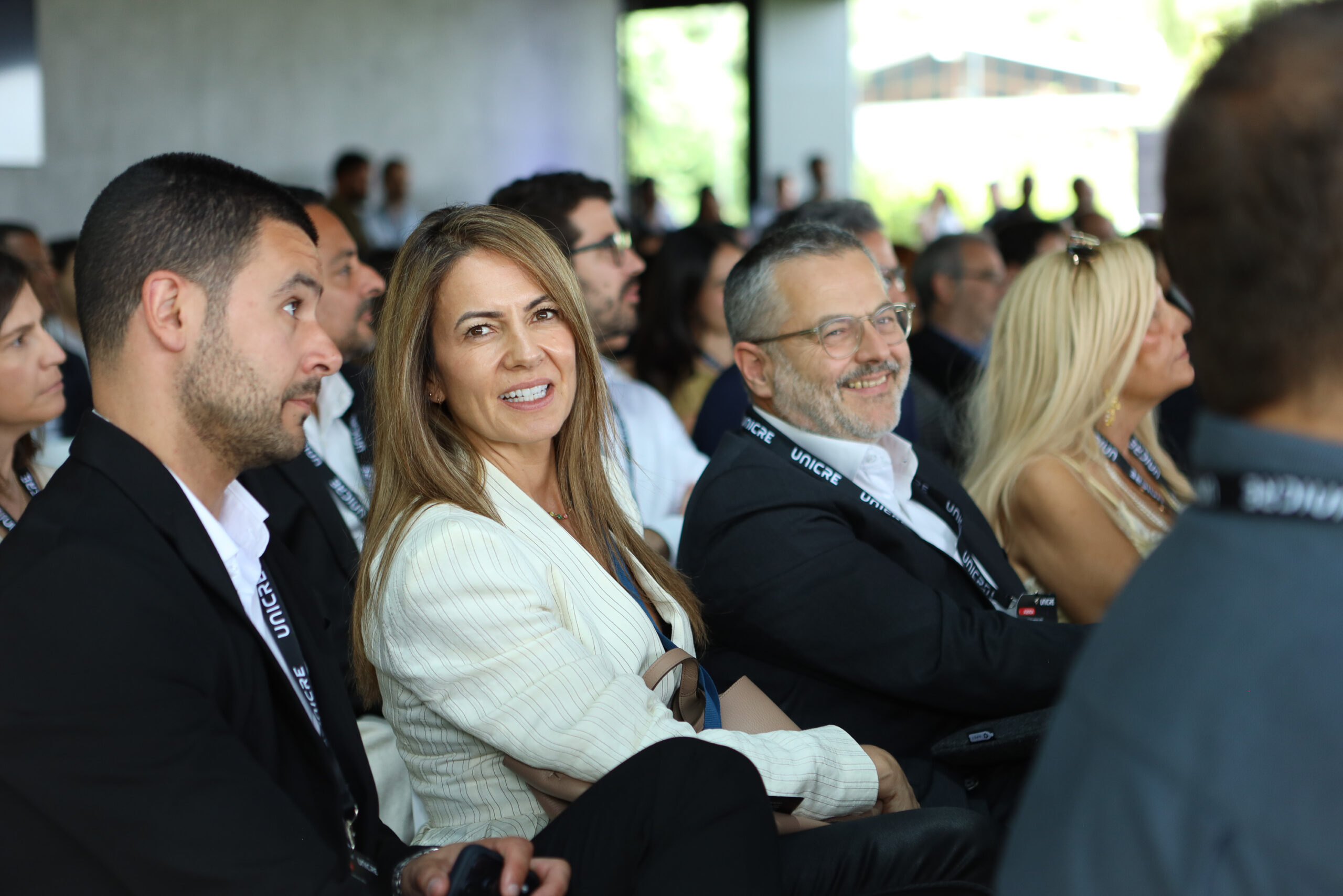 Smiling woman in a striped blazer seated among conference attendees with a lanyard.