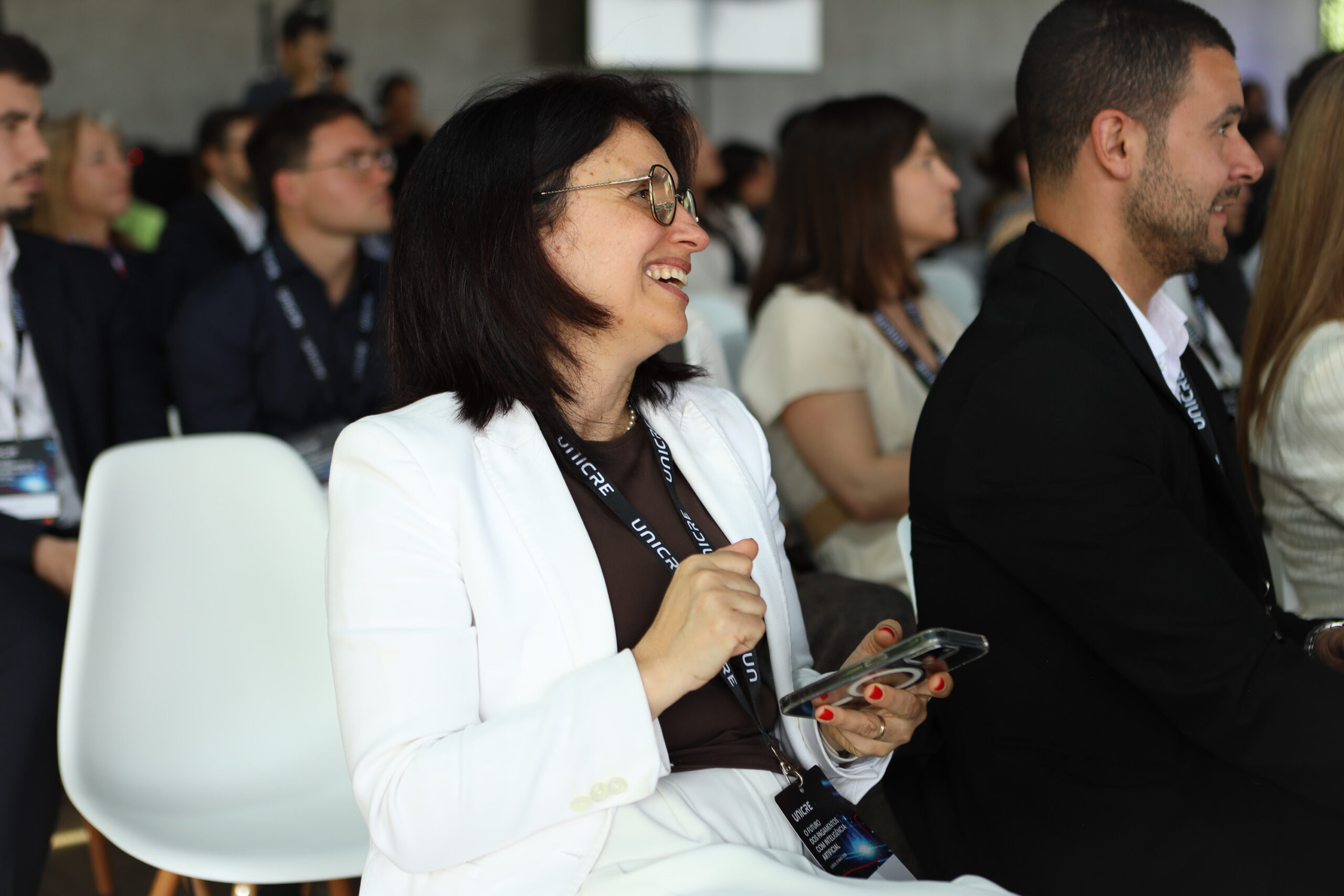 Smiling woman with glasses in a white blazer at a conference, holding a phone, with other attendees in the background.