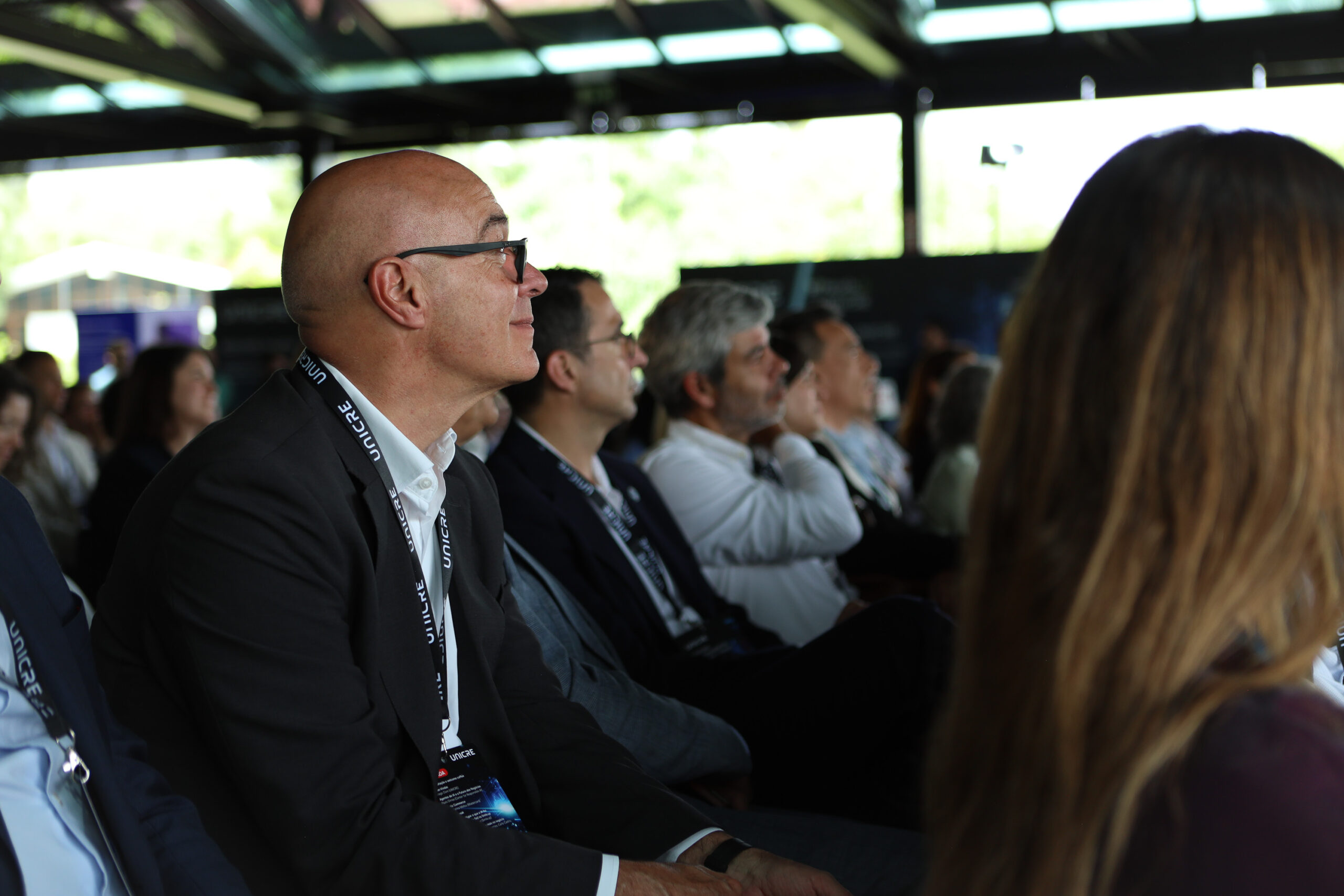 Side profile of a bald man wearing glasses and a lanyard, seated in a conference audience under a covered structure.