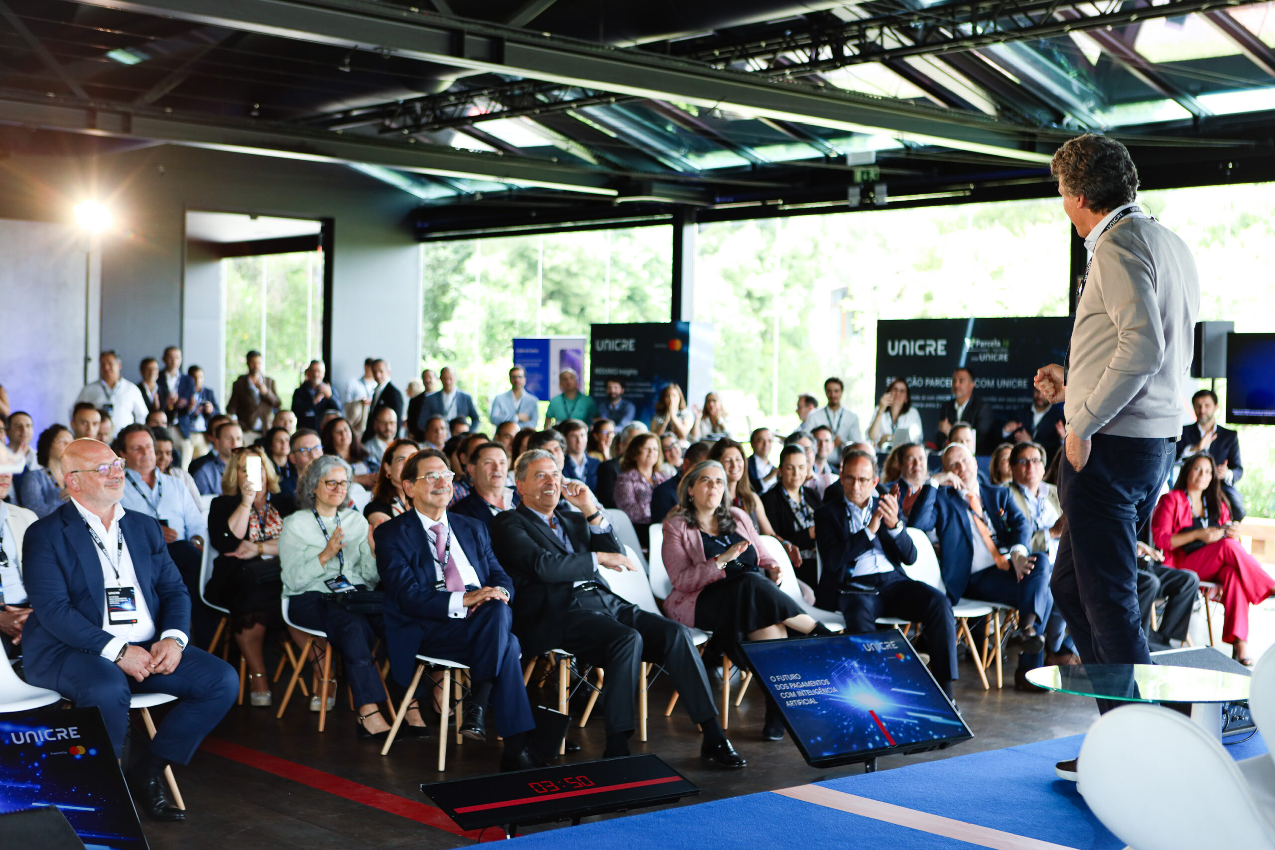 Speaker presenting to a large seated audience at a conference, with UNICRE banners in the background.