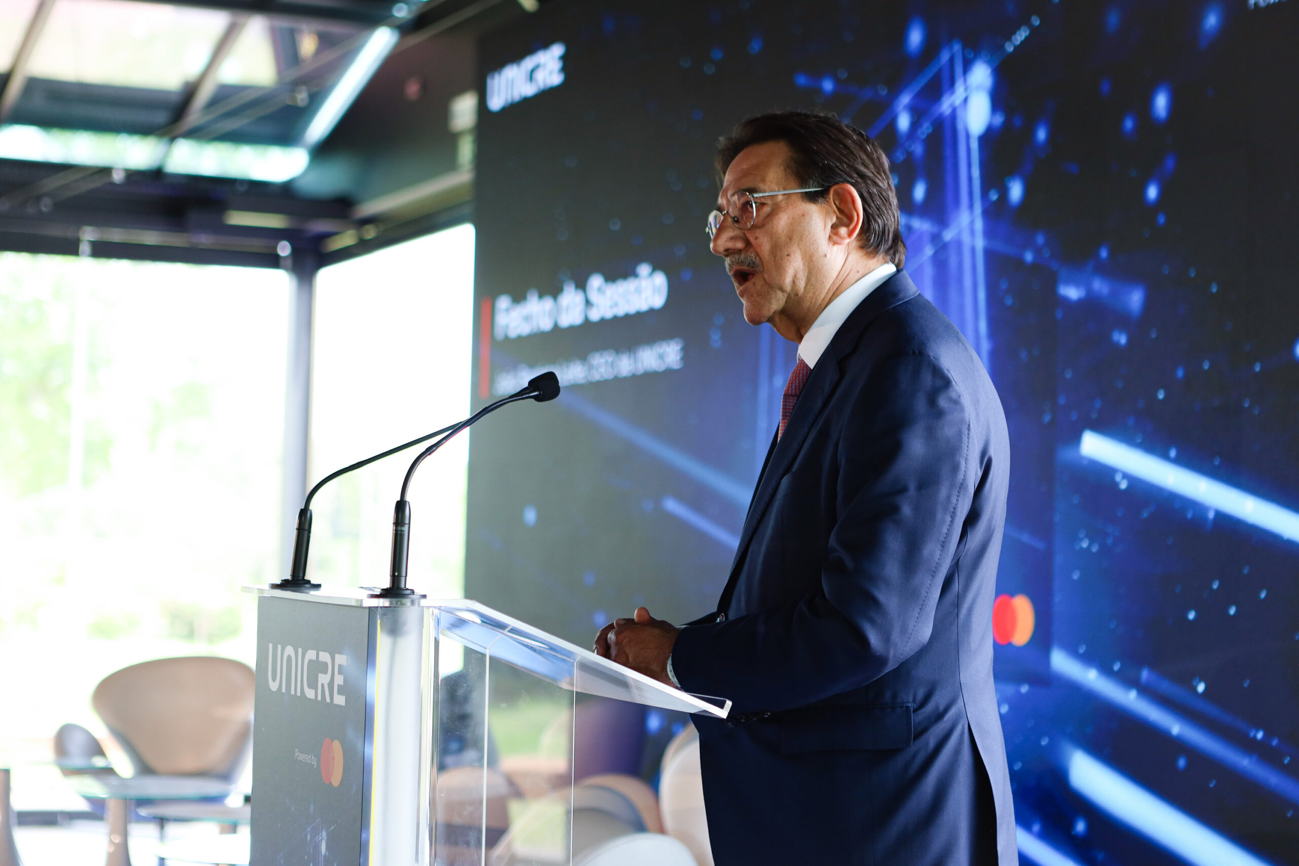 Man in a dark suit and glasses speaks at a transparent podium with two microphones, a blue tech-themed backdrop behind him.