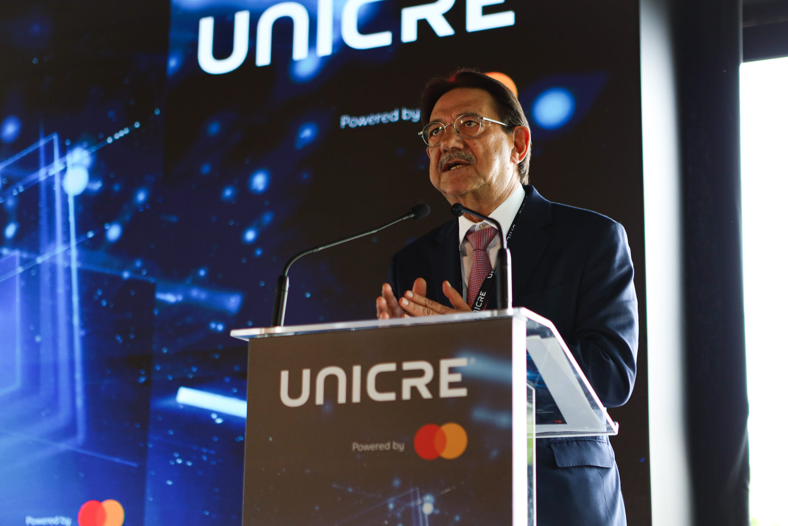 Man in a suit and glasses delivering a speech at a podium with a UNICRE backdrop and Mastercard logo visible on the banner behind him.