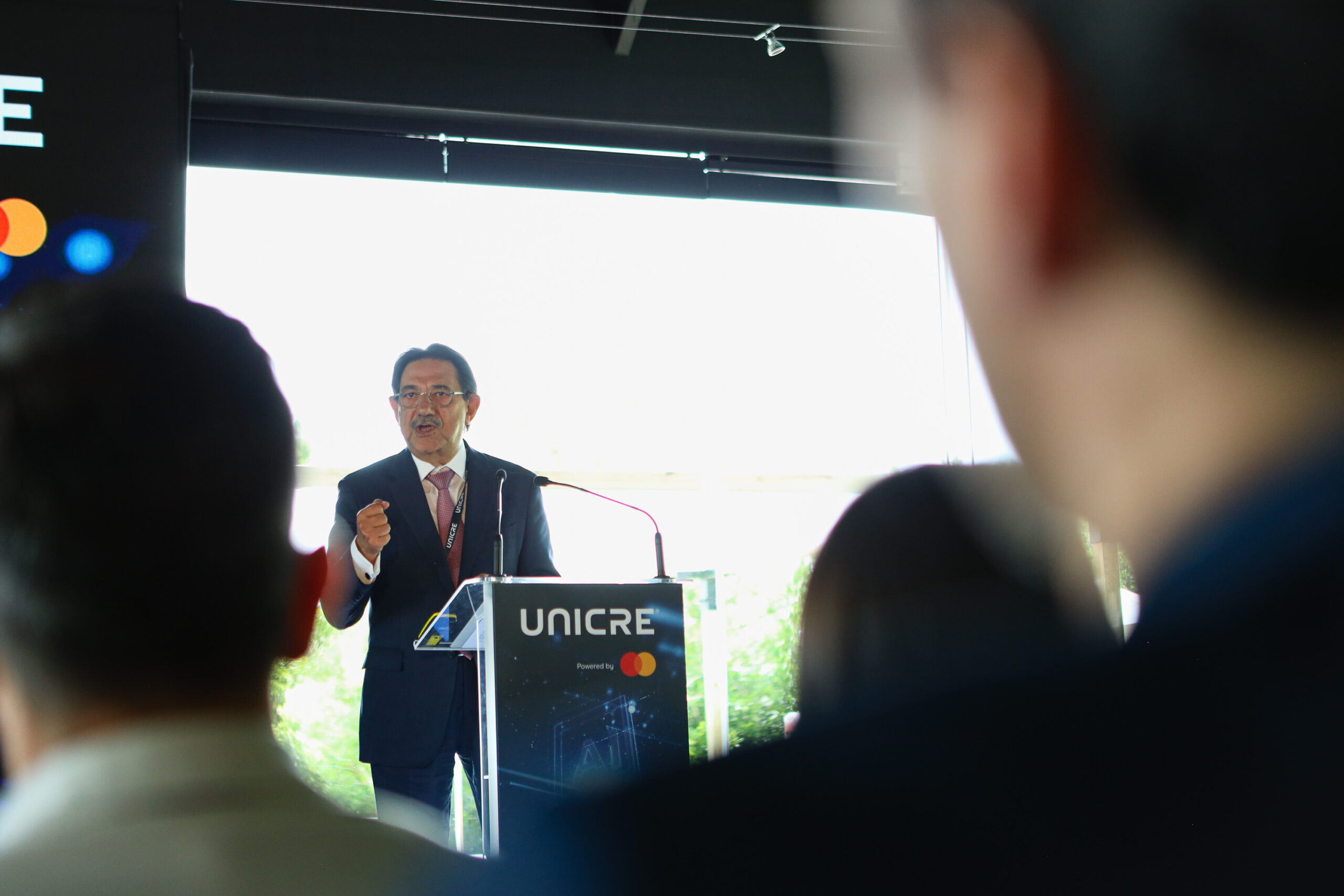 Speaker in a suit delivering a talk at a UNICRE-branded lectern, audience silhouettes visible in the foreground.