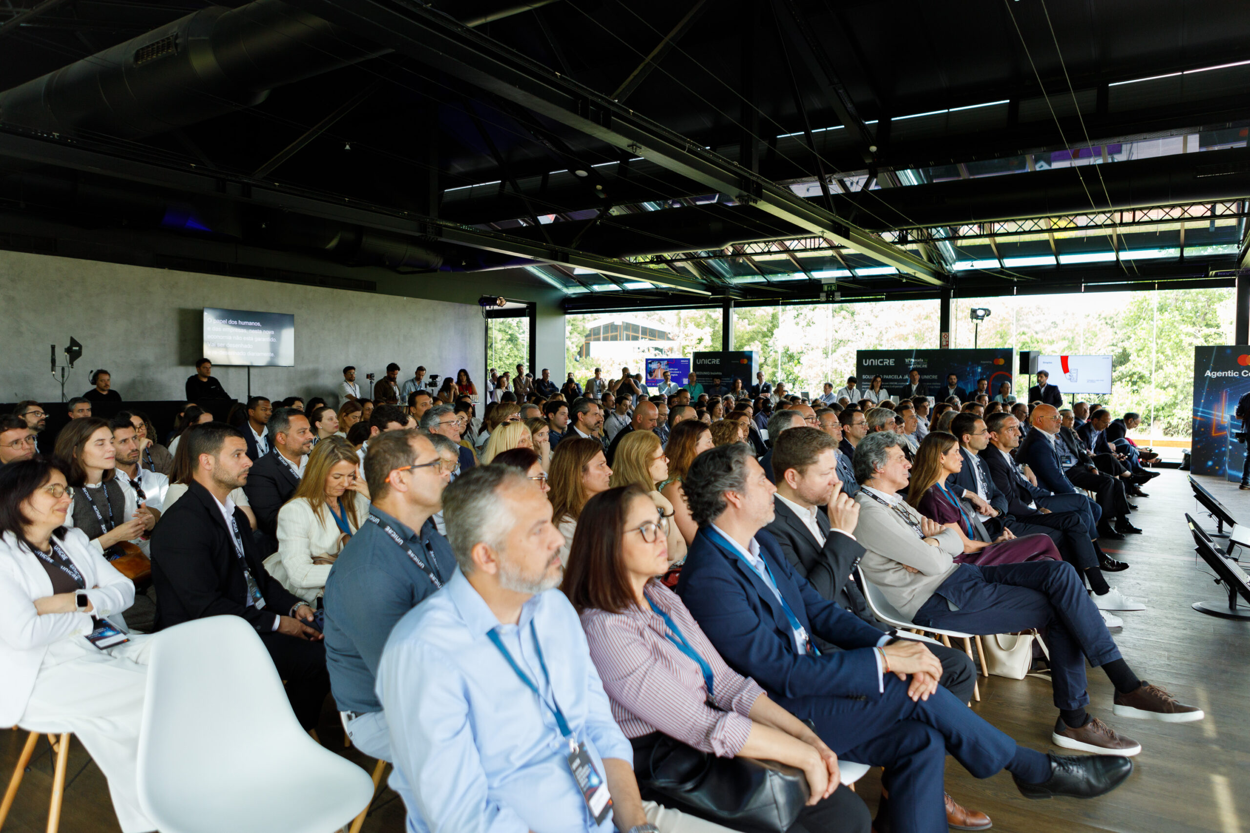 Audience seated in a large conference hall watching a presentation, with banners at the back and natural light outside.