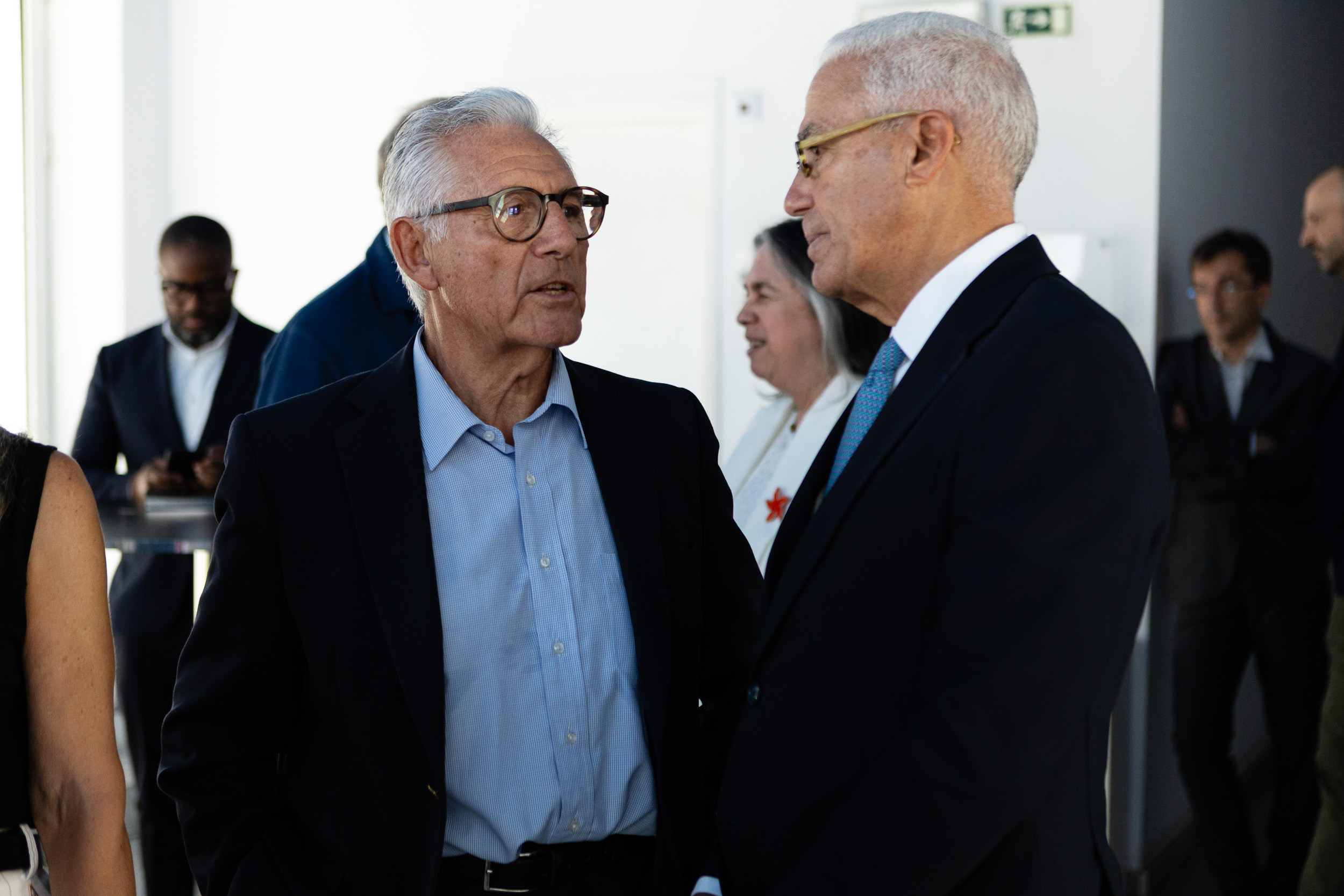 Two suited men in a professional conversation at a networking event, facing each other and speaking, with others in the background.