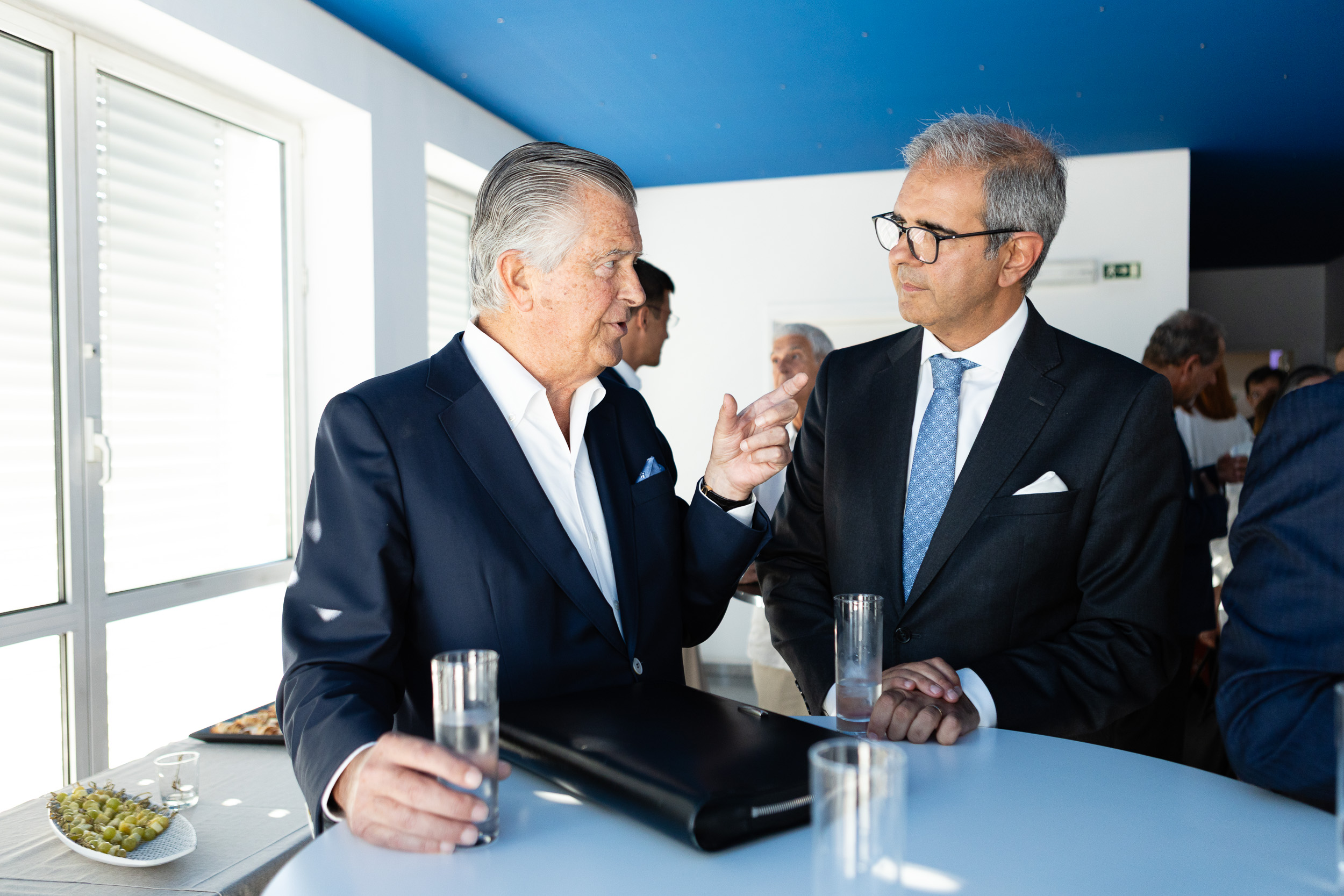 Two older businessmen in dark suits chat at a sleek blue-toned counter, one gesturing with a finger while holding a glass of water; snacks in view nearby.