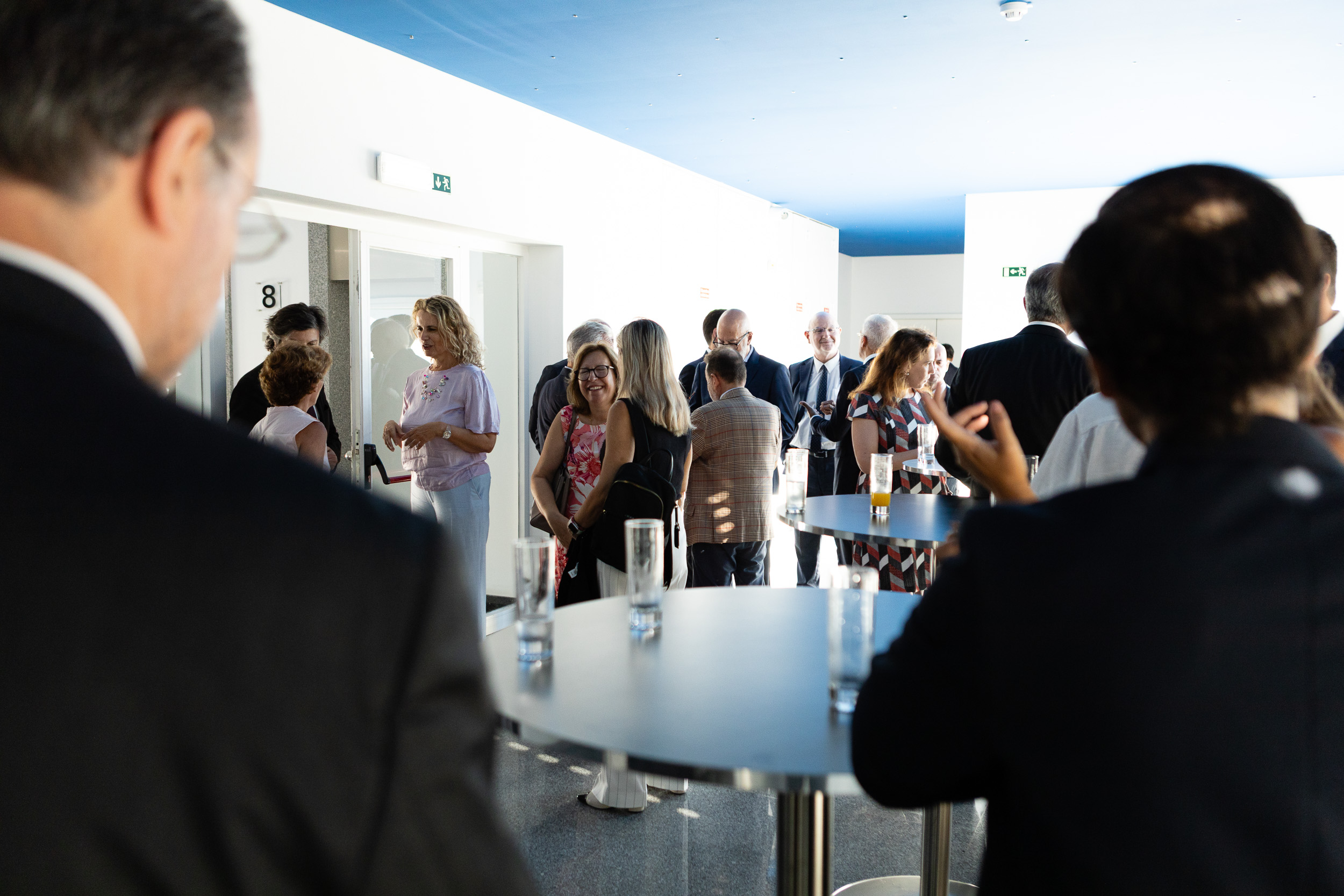 People mingling at a casual indoor reception, standing around high tables with drinks while conversing near white walls and a blue ceiling.