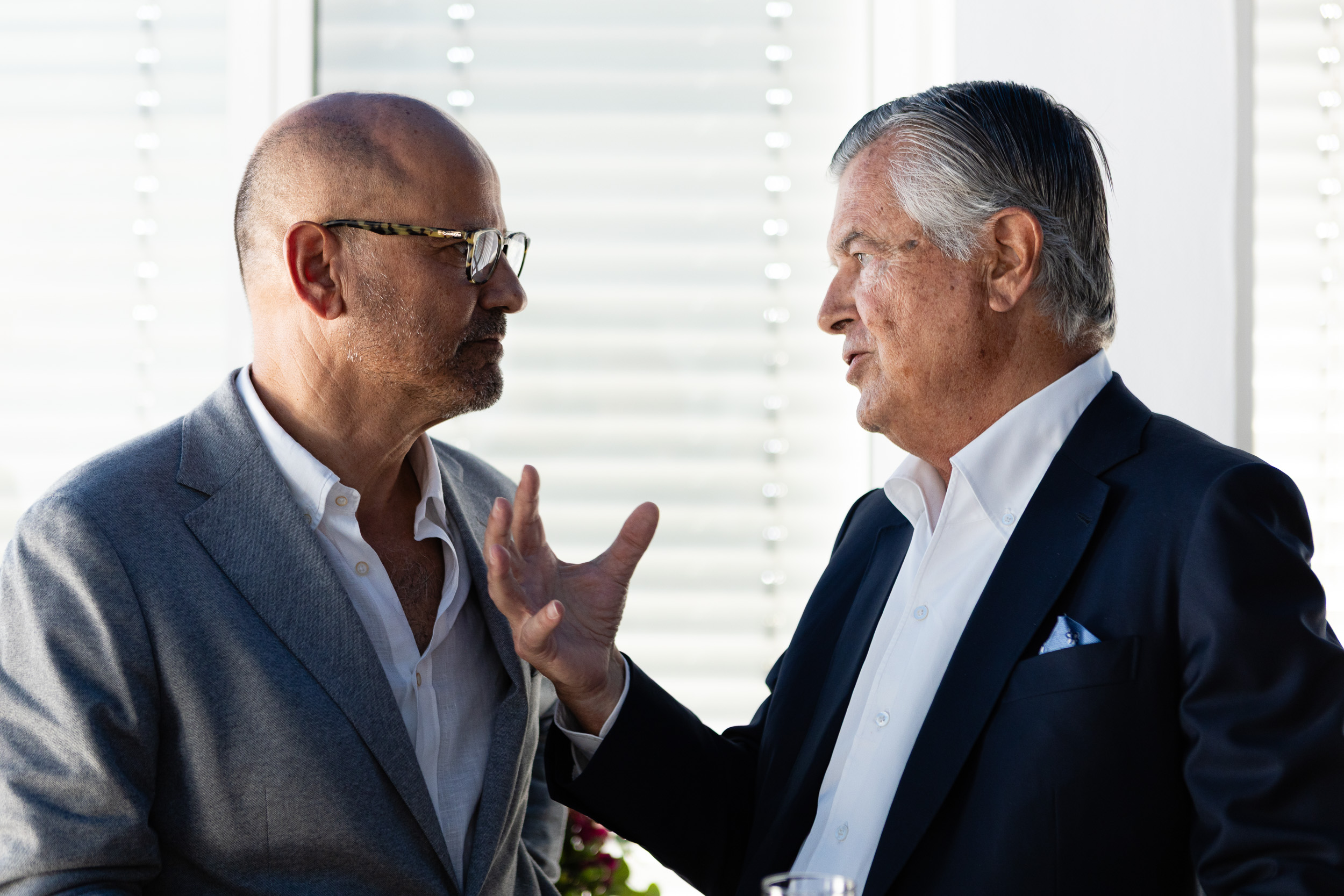 Two middle-aged men in business suits converse closely, one gesturing with his hands.