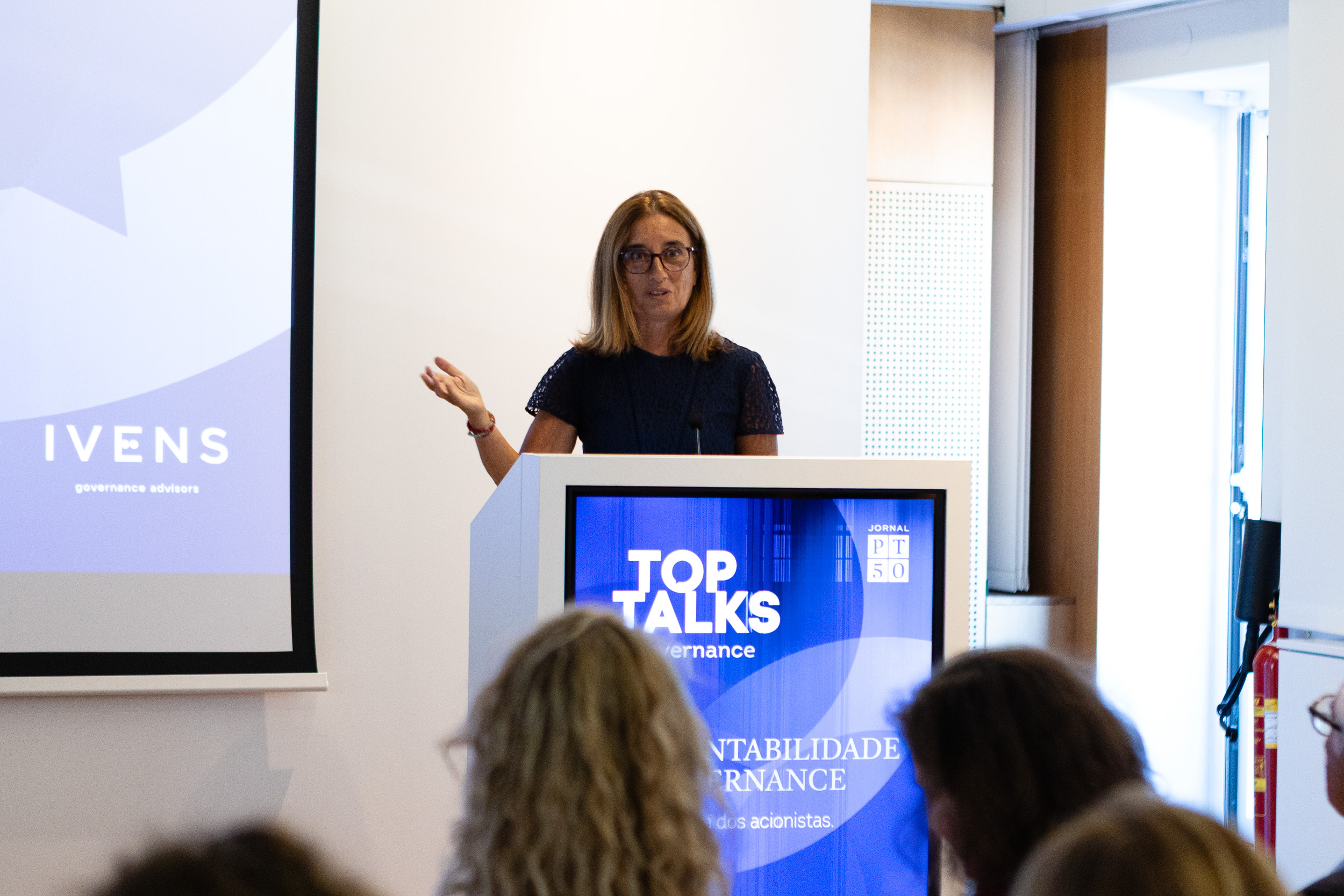 Female speaker at a podium giving a talk, audience in foreground, screen reads 'TOP TALKS' and 'Governance' in blue.