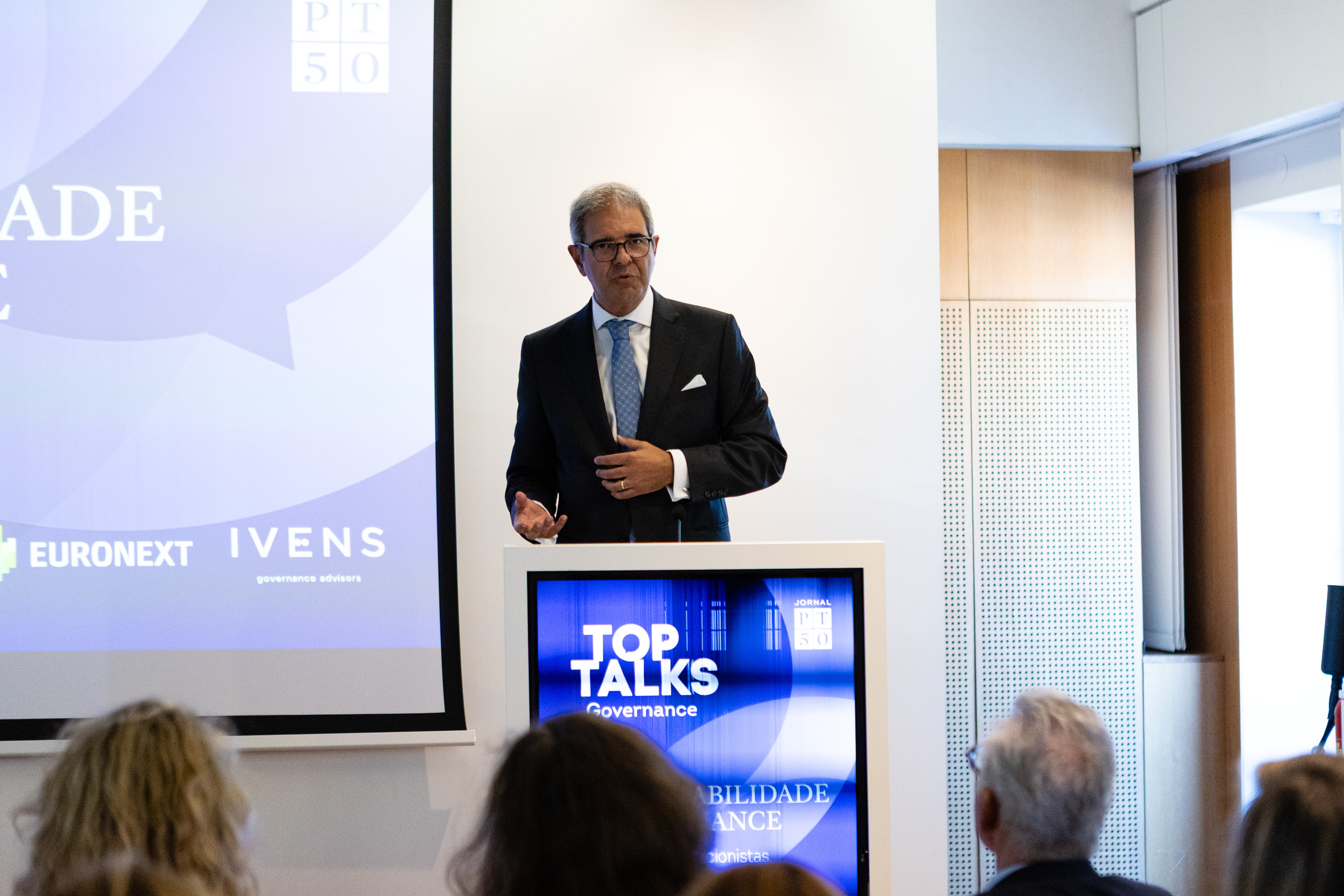 Male presenter in a dark suit and blue tie speaks at a podium in a conference room, with screens showing 'Top Talks' and governance behind him, audience in foreground.