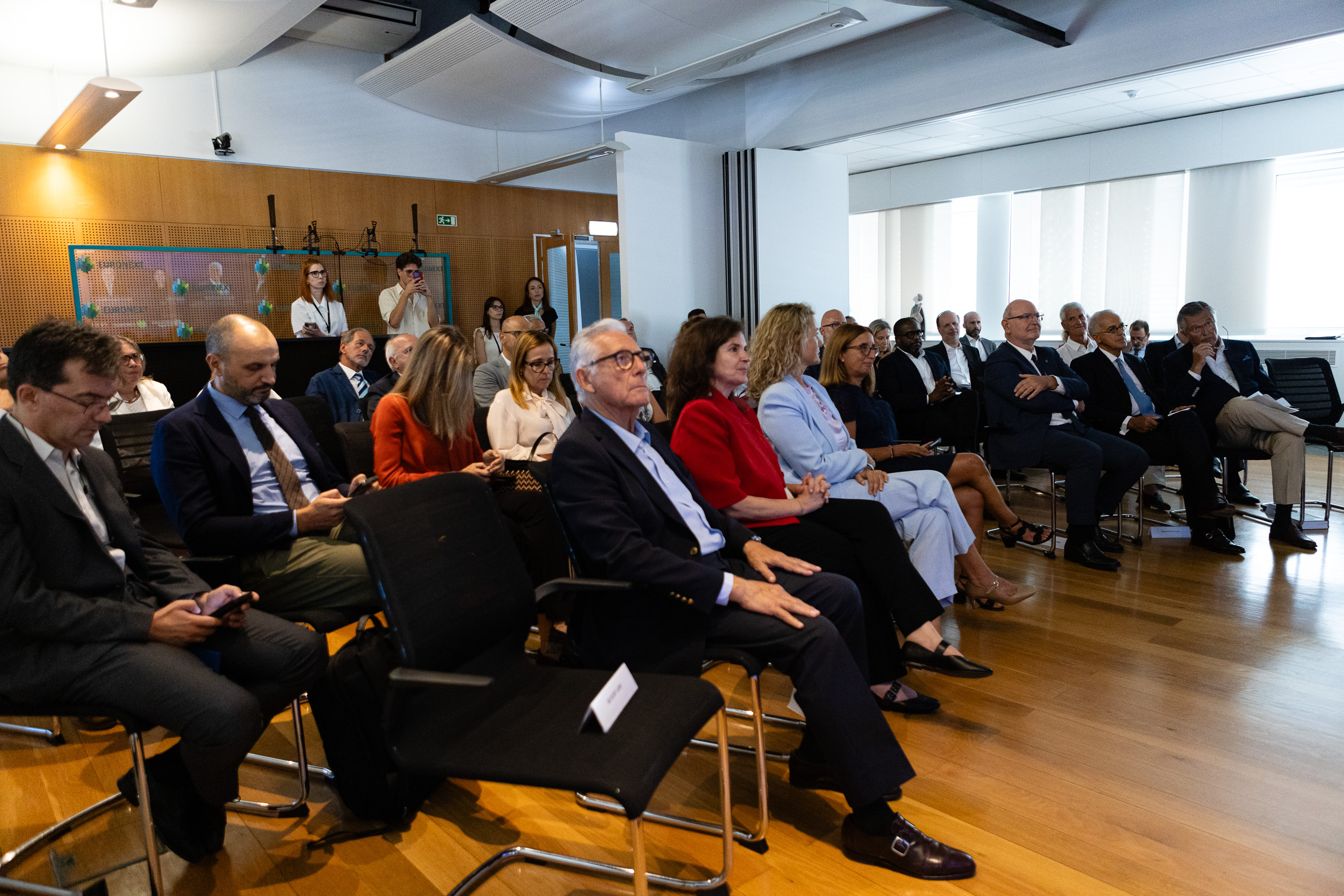 Group of professionally dressed attendees seated in a conference room, attentively listening to a presentation on a wooden-floored venue.