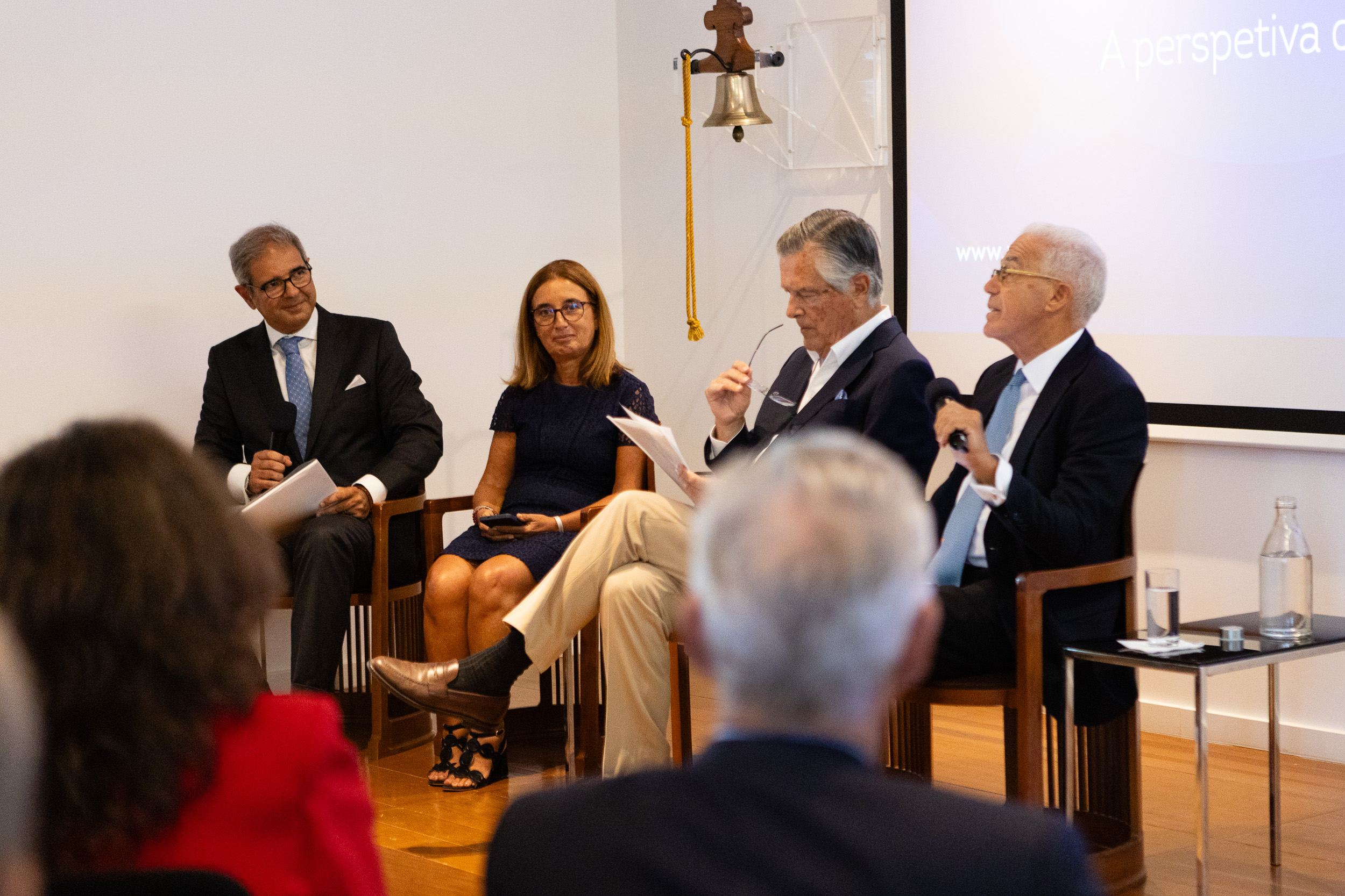 Panel discussion with four speakers seated in a row, audience in foreground, man with microphone on left, woman in blue dress, two men on the right; projection screen in background.