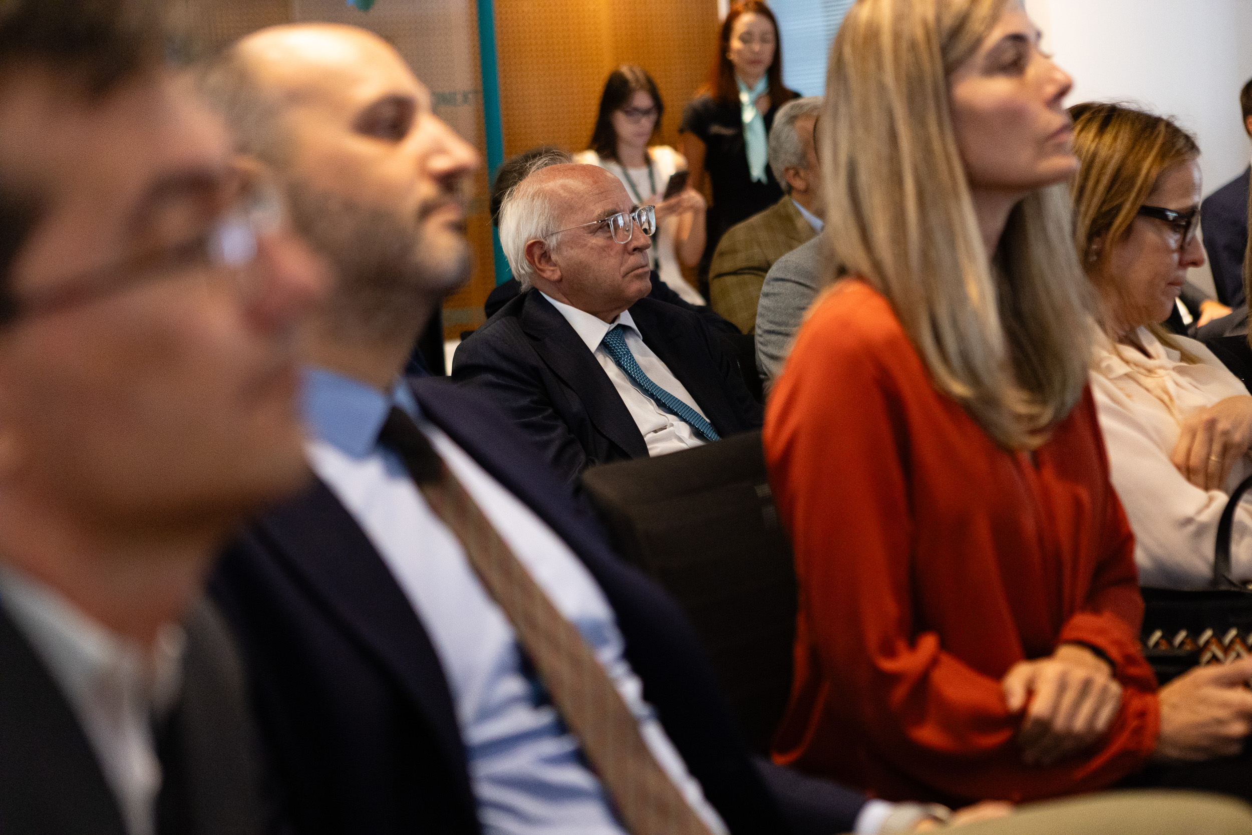Audience at a conference: a white-haired man in glasses and a suit sits center, others around him listening.