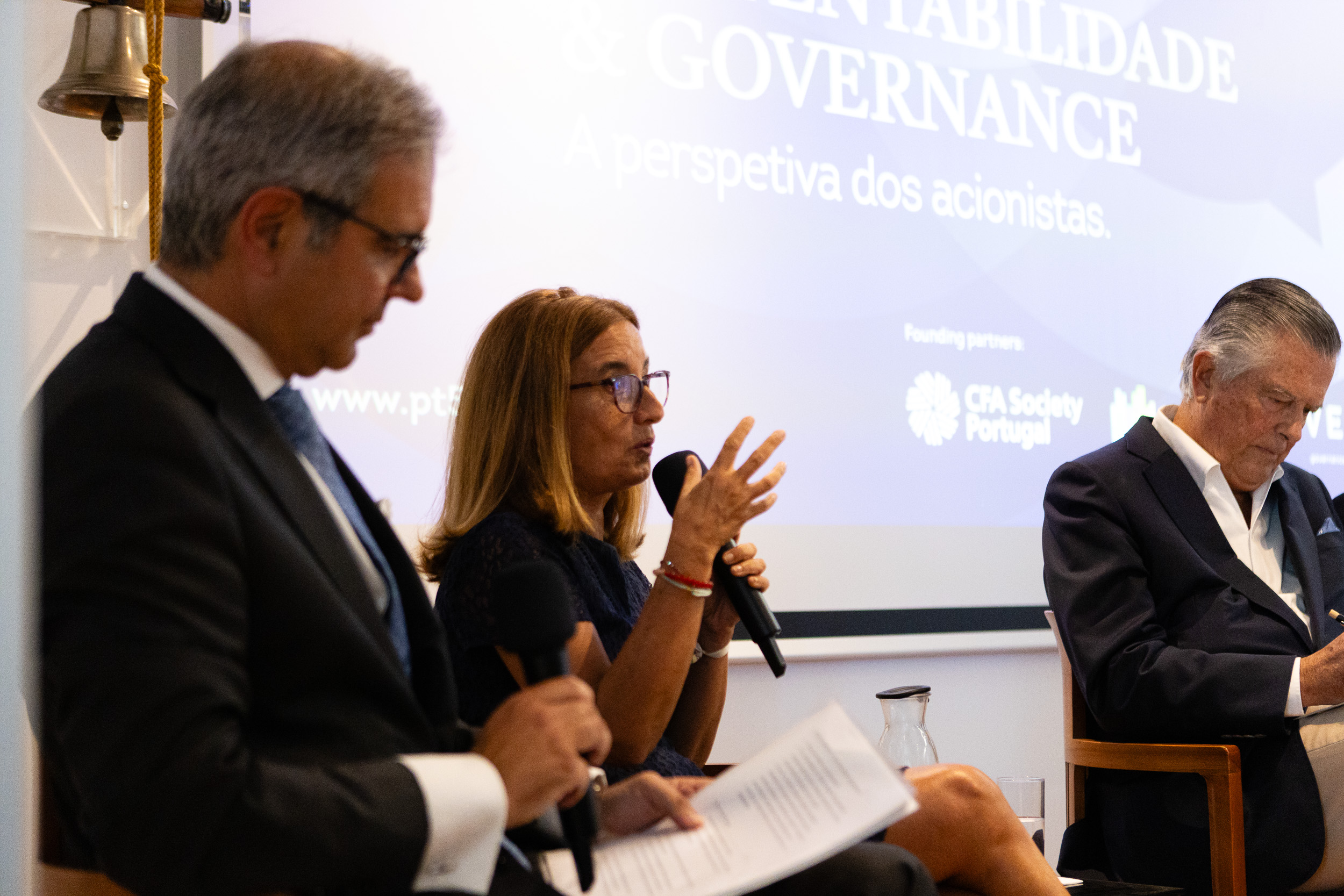 Panel of three speakers in a conference room; woman center speaks into a microphone while others read or listen, a projection screen behind them.