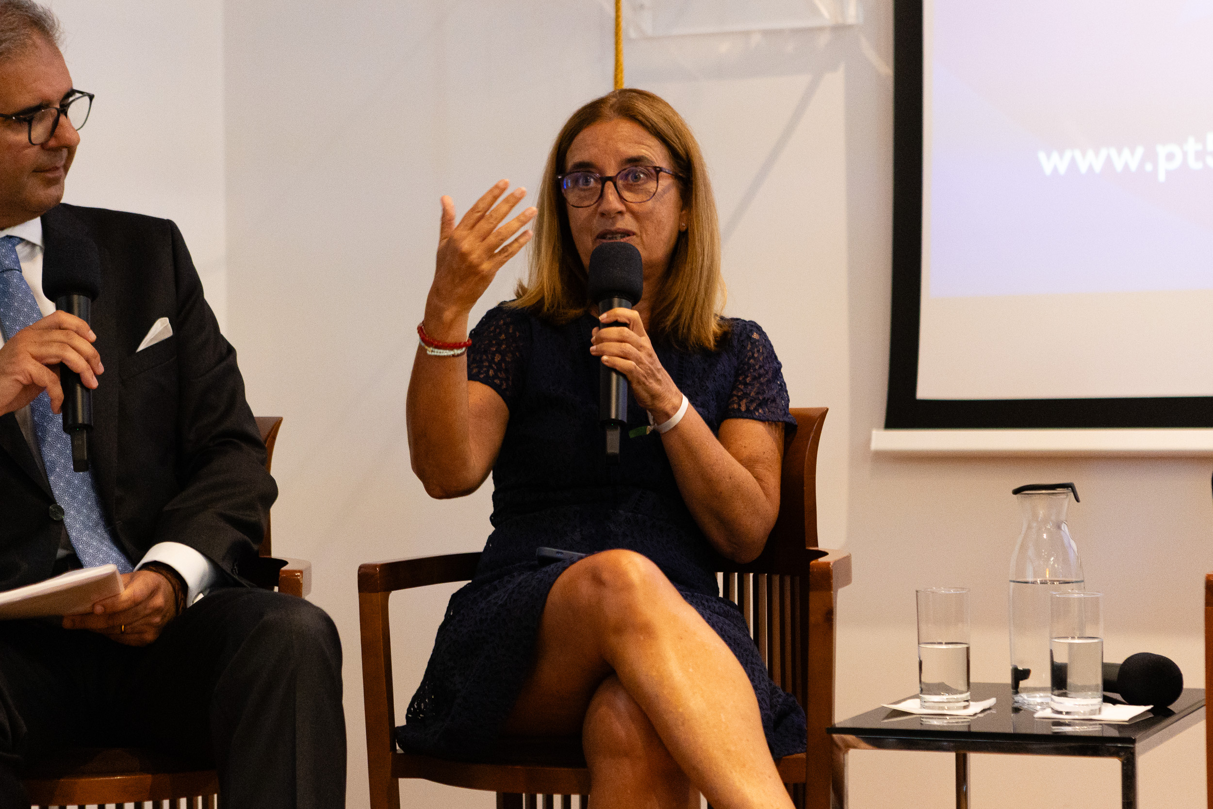 Woman panelist in a navy lace dress speaks into a handheld microphone, gesturing with her left hand while seated beside a man in a suit during a discussion, with water glasses on the table nearby.