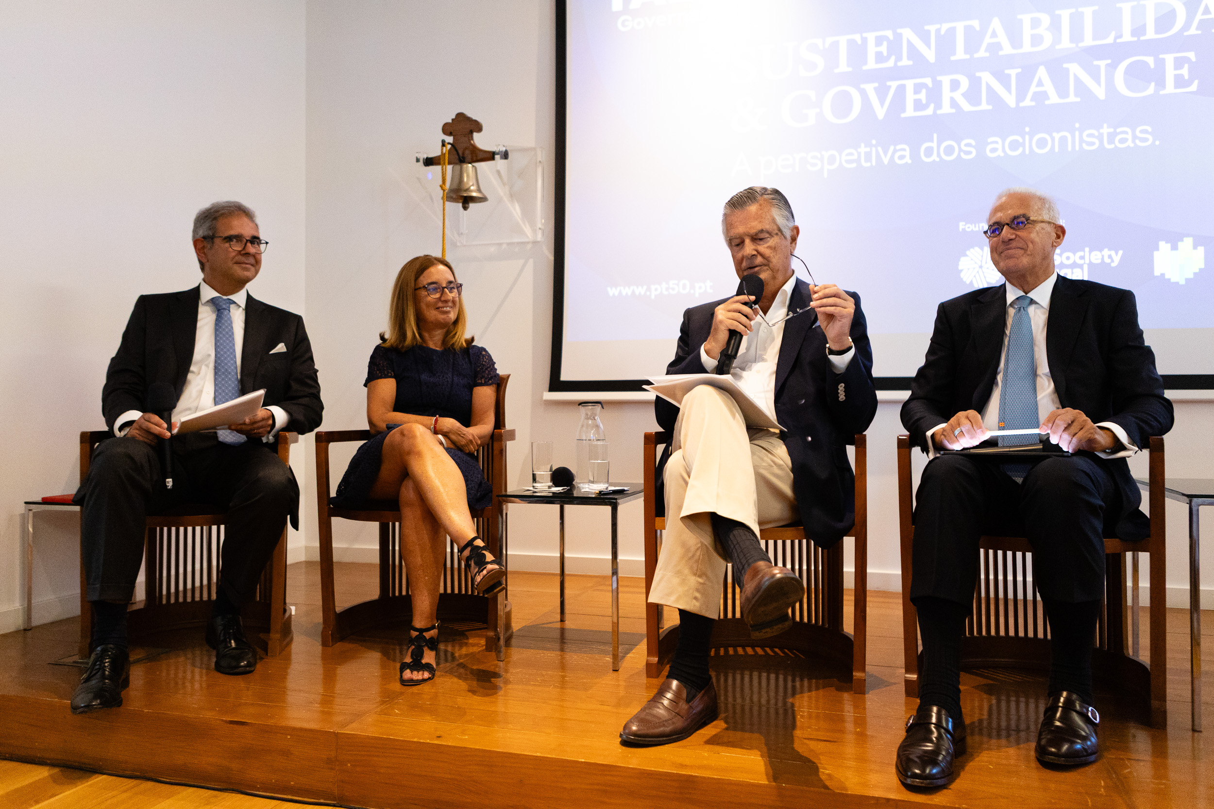 Panel of four professionals in suits seated on stage during a discussion, with a projection screen behind them displaying text.