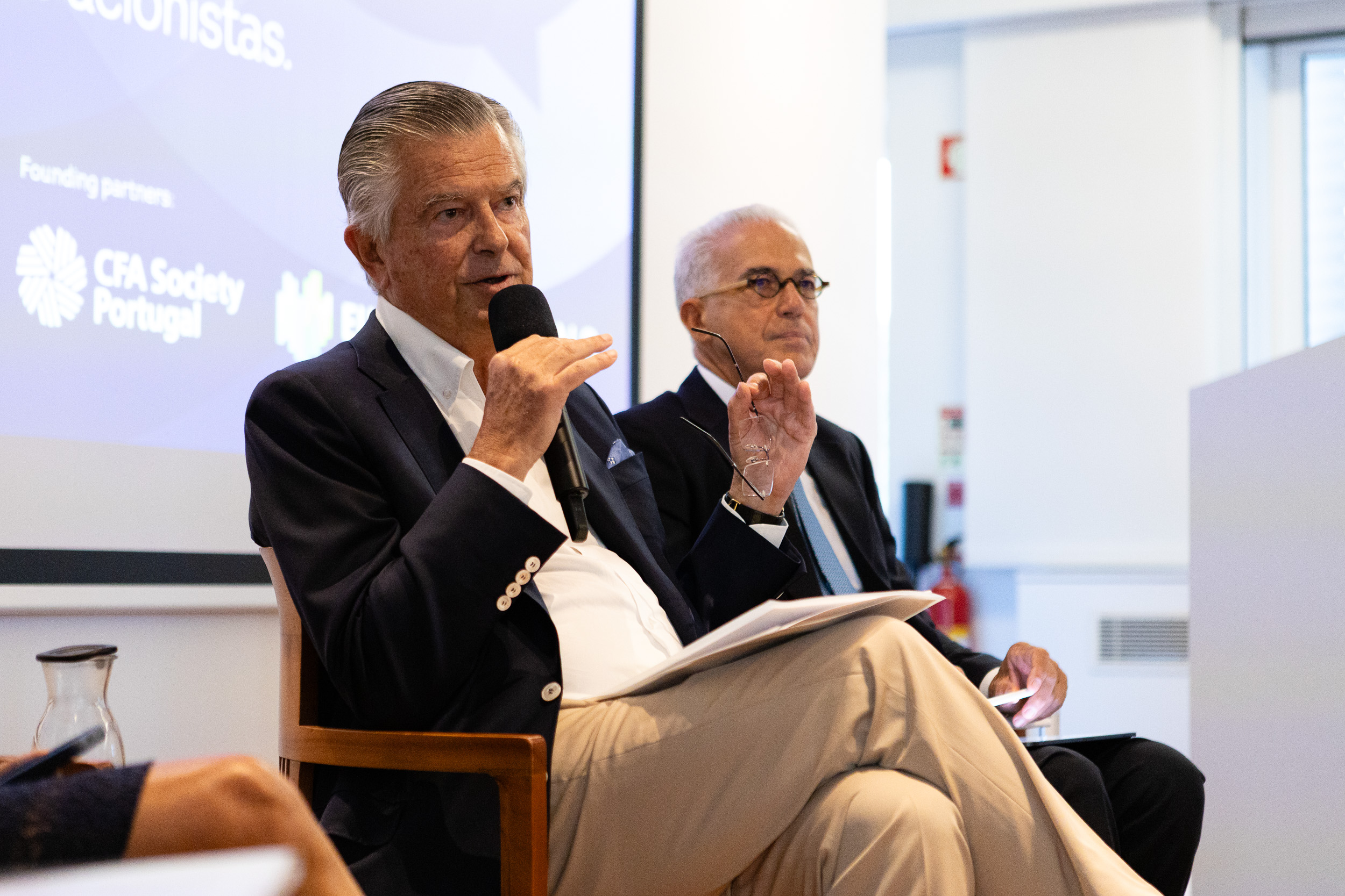 Two men in suits seated on a panel; one speaks into a handheld microphone, background displays CFA Society Portugal text?
