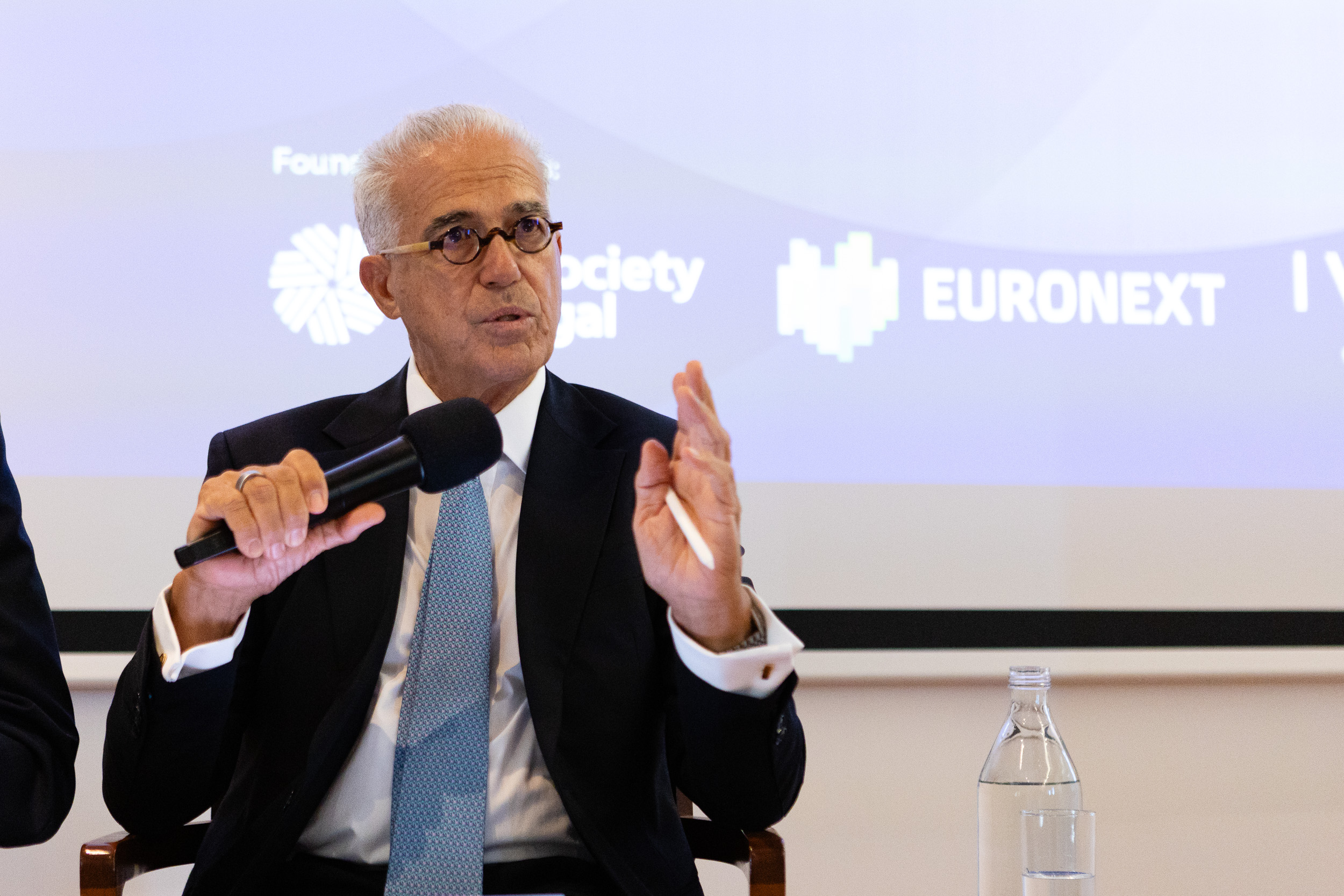 Older man in a dark suit and round glasses speaks into a handheld microphone at a panel, with EURONEXT branding in the blue background.