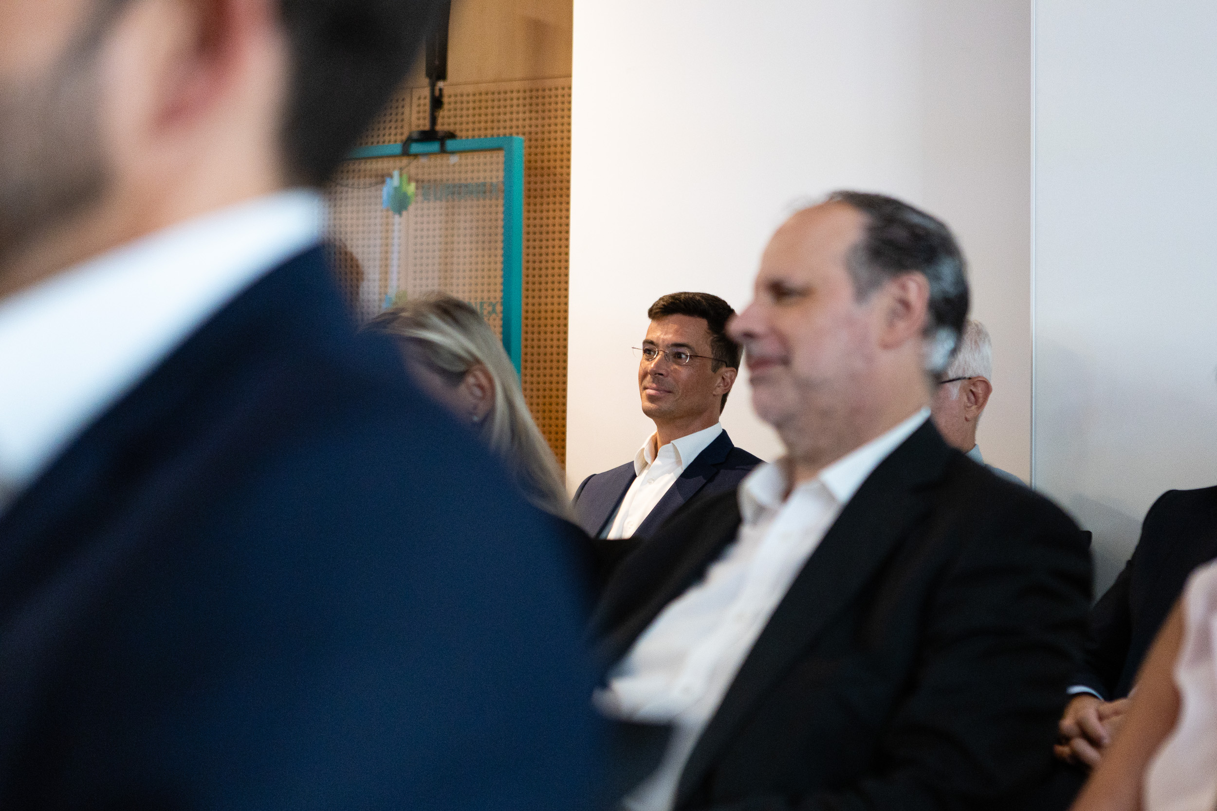 Professional meeting scene with a man wearing glasses seated and looking to the left, others blurred in the foreground.