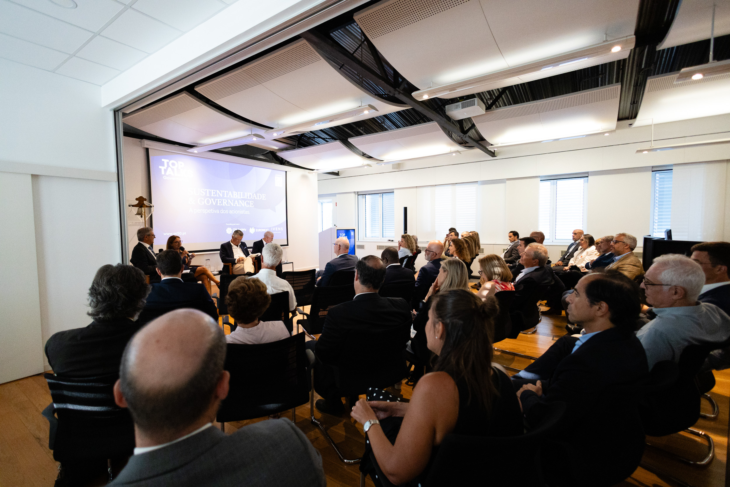 Audience seated in a bright conference room watching a panel discussion on stage with a large projection screen behind them