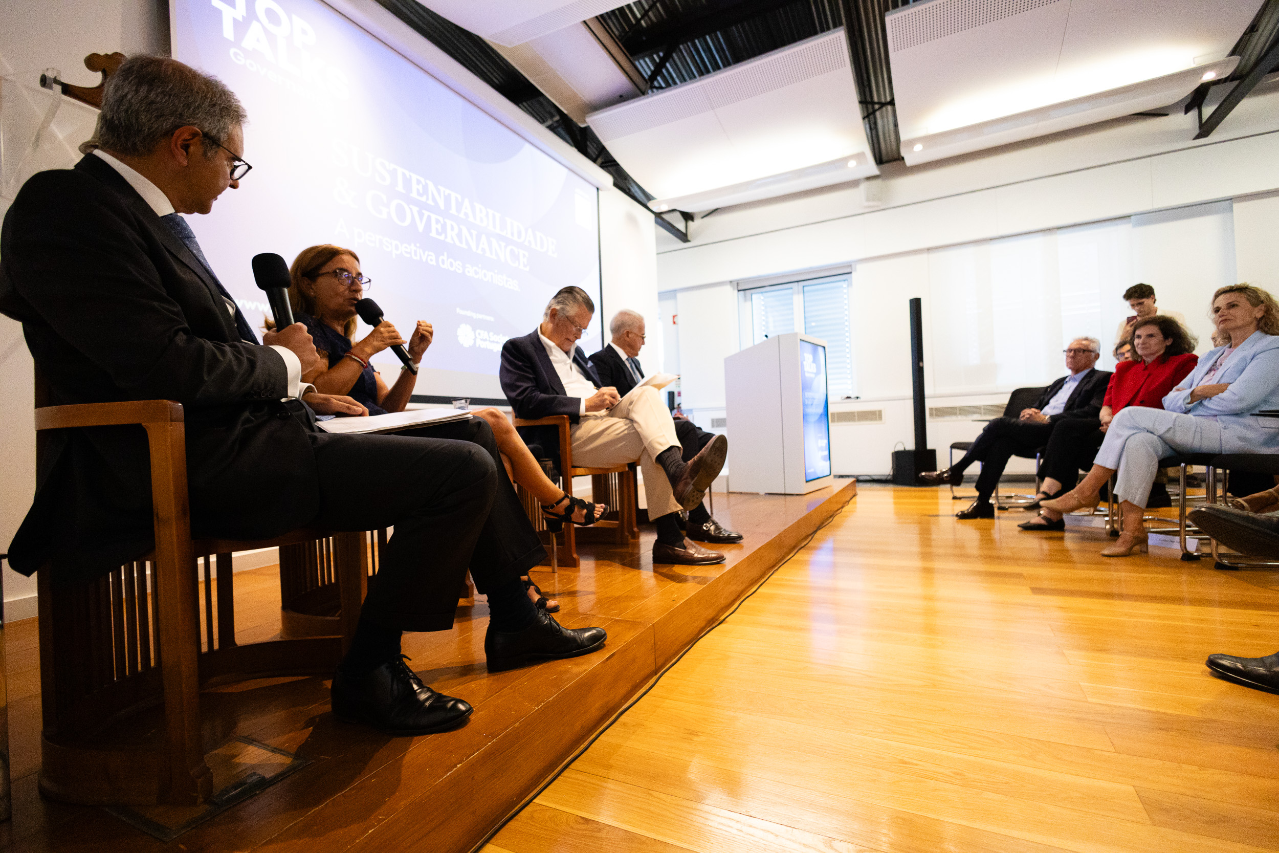 Panel discussion on a stage in a bright conference room; four panelists with microphones, audience seated to the right, projection reads 'SUSTENTABILIDADE & GOVERNANÇA'.