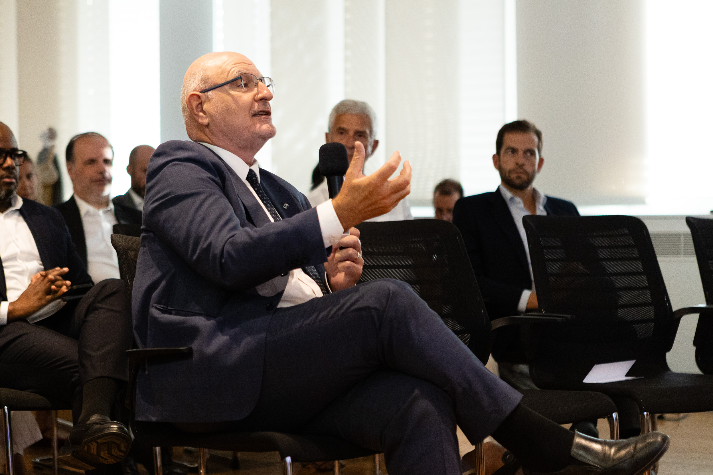Man in a blue suit speaking into a handheld microphone during a panel discussion in a conference room.