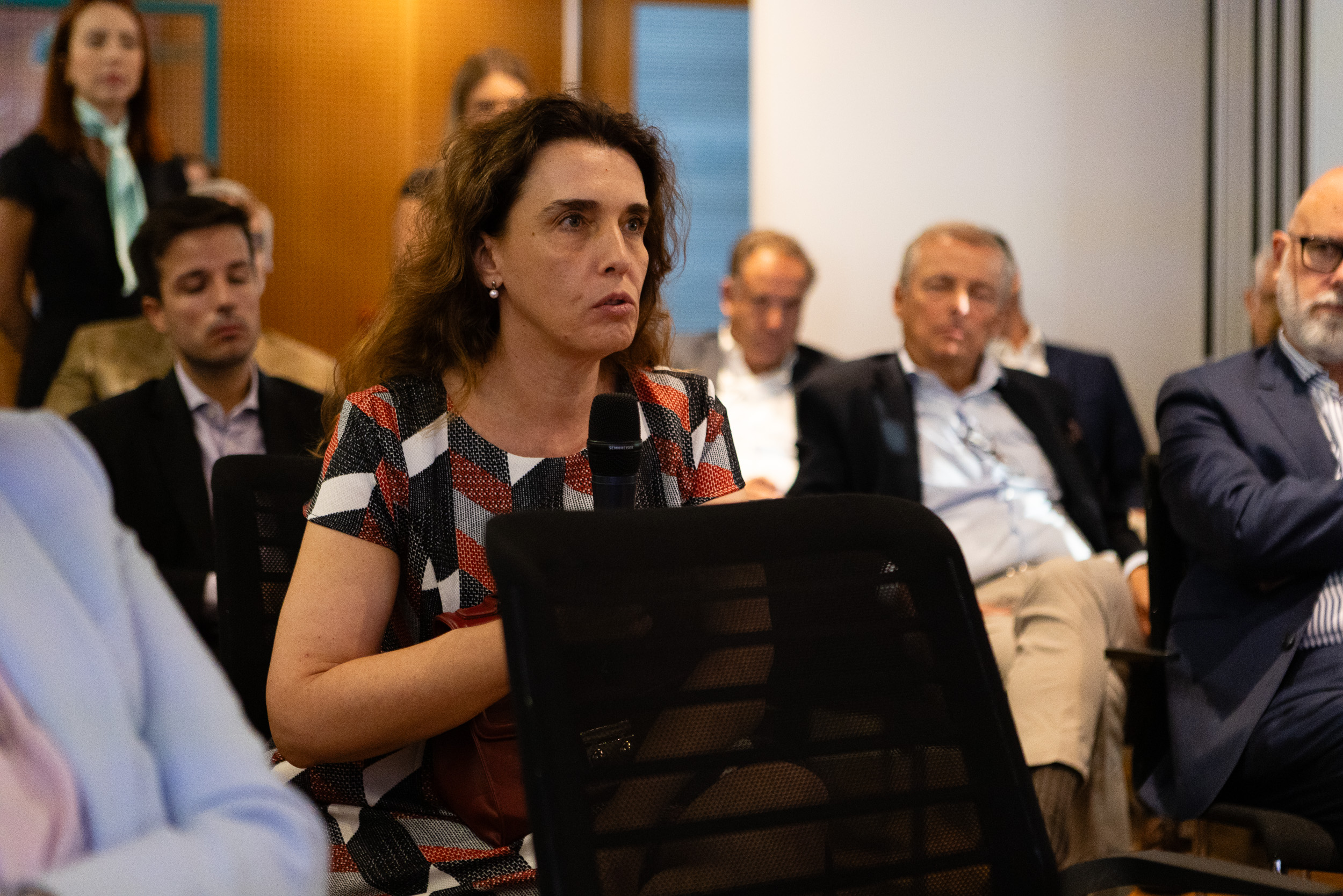 Woman in a patterned dress speaks into a handheld microphone at a conference, surrounded by seated attendees.
