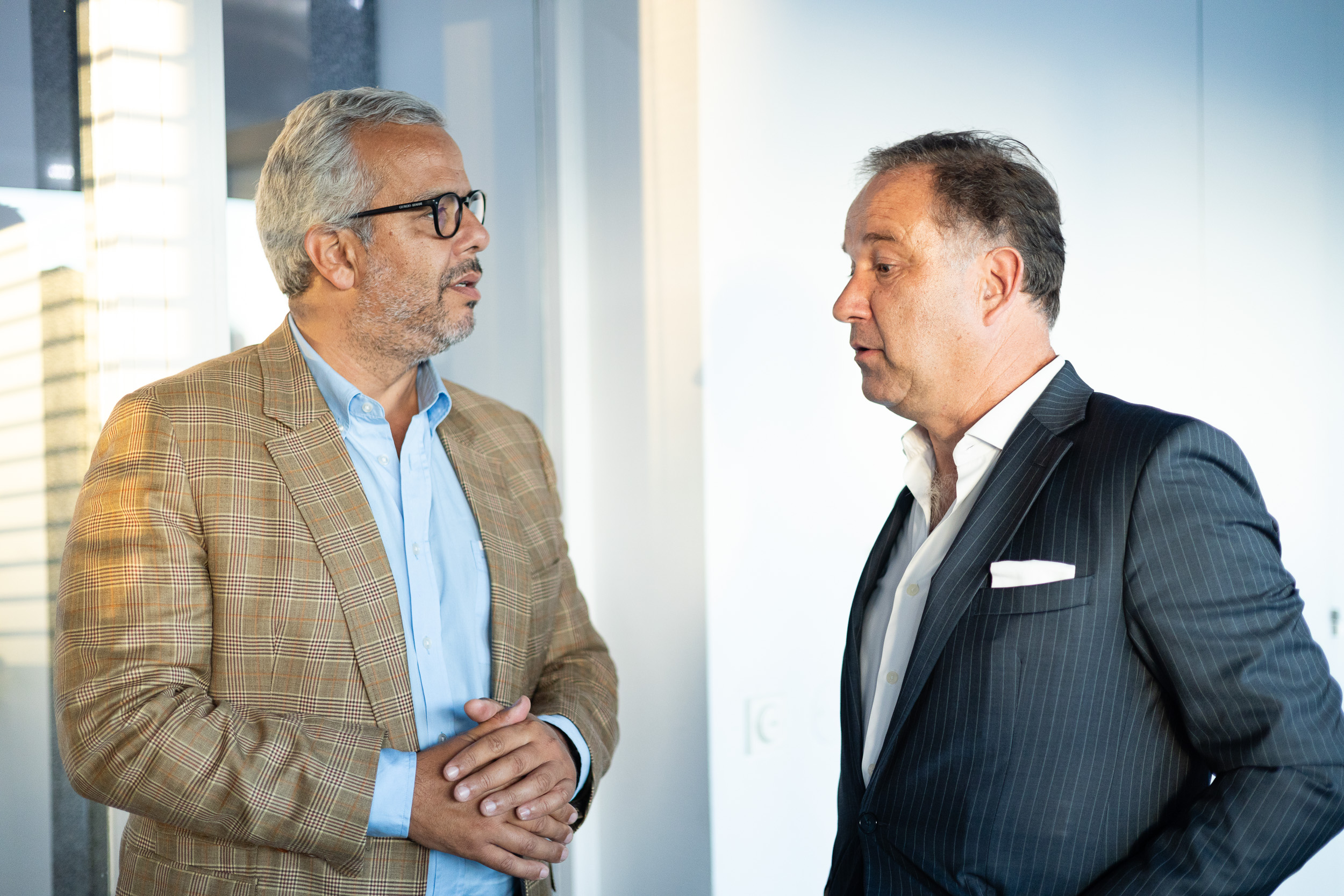 Two middle-aged businessmen in an office doorway facing each other, with hands clasped in a handshake as they talk.