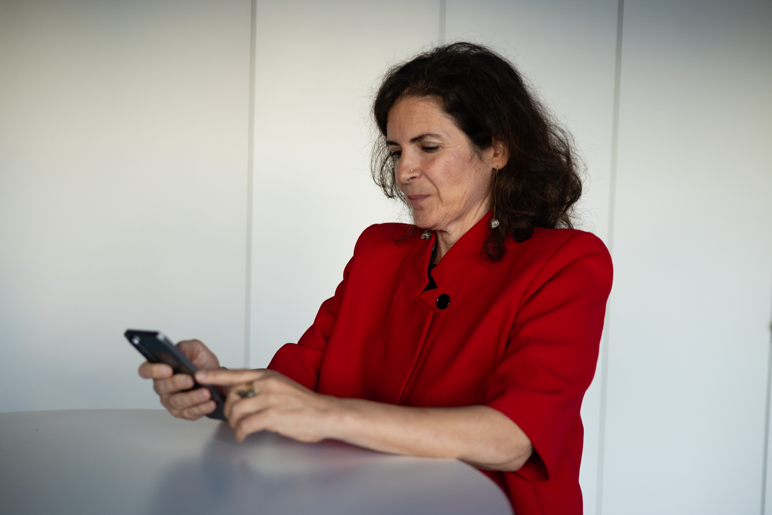 Woman in a red jacket sits at a table, focused on a smartphone in her hands.
